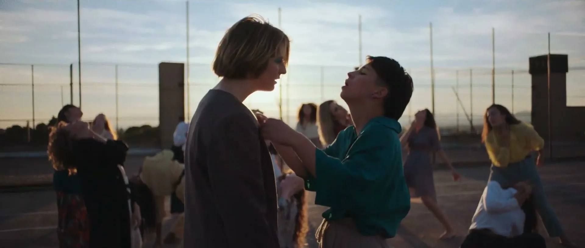 Two young women, one with short hair and one with long hair, embrace and look up at the sky on an outdoor court with other people in the background.