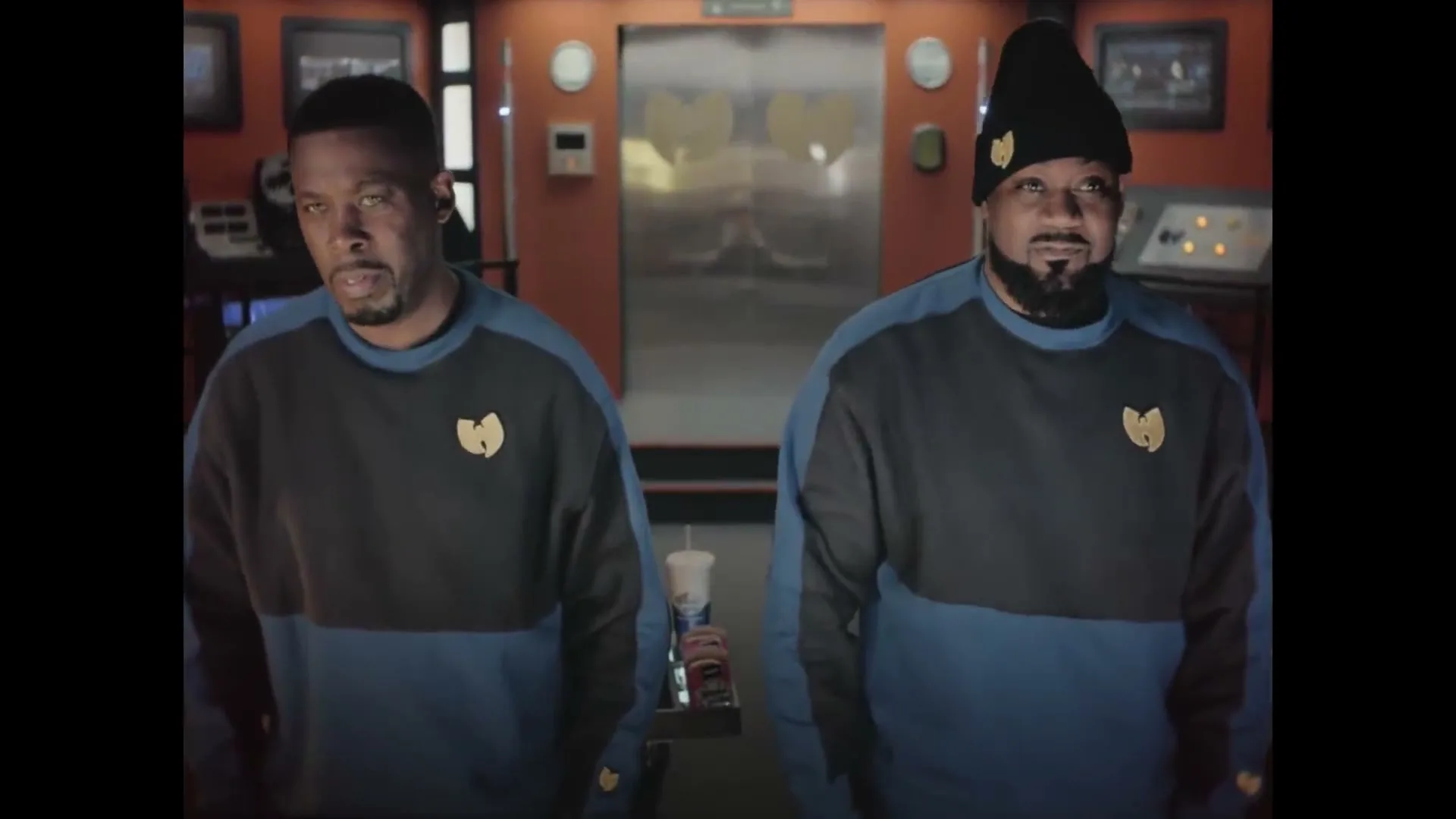 Two men in Wu-Tang Clan branded sweaters stand in a spaceship control room, looking around with curious expressions.