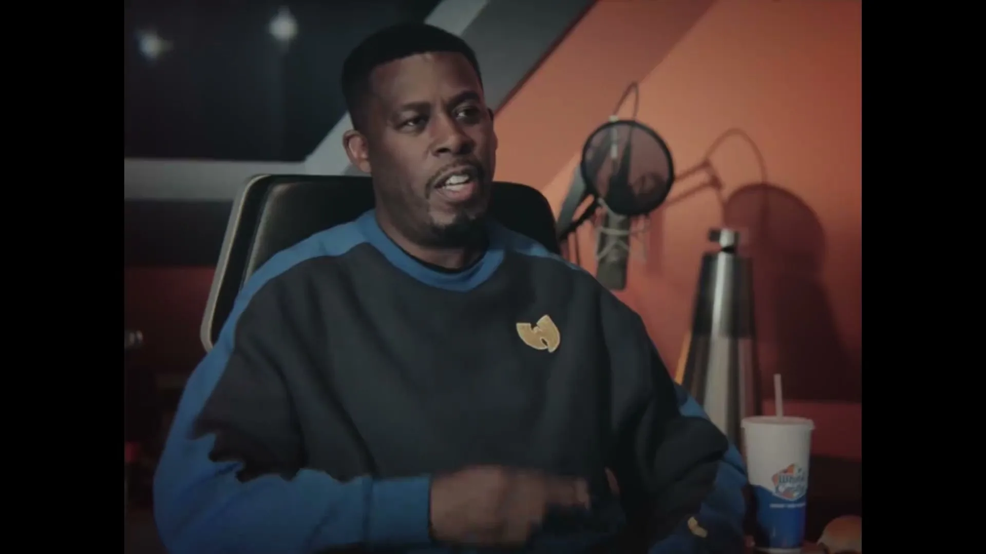 A man in his 40s, wearing a blue and black sweatshirt with a Wu-Tang Clan logo, sits in a recording studio, talking.