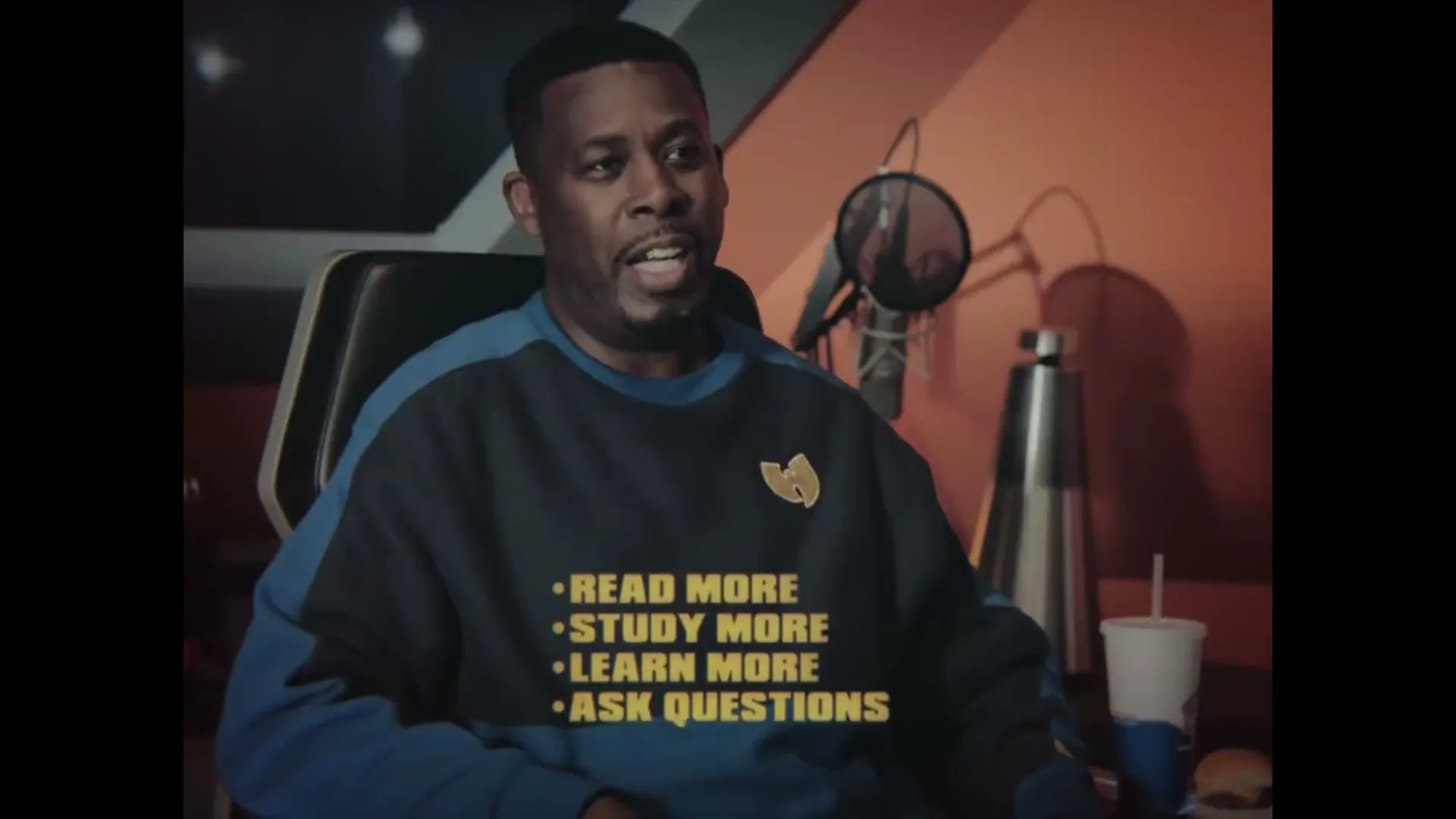 A Black man in his 40s, wearing a blue and black Wu-Tang Clan sweatshirt, sits in a recording studio, speaking with a serious expression.