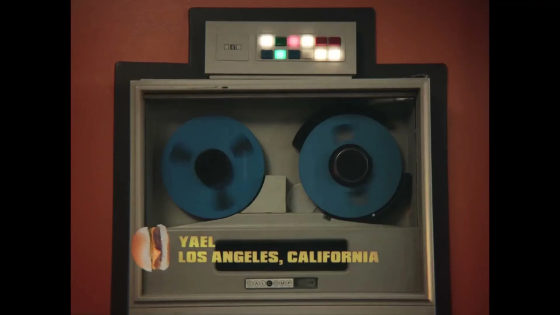 A vintage reel-to-reel tape recorder with blue reels spins, displaying the name "YAEL" and "LOS ANGELES, CALIFORNIA" in yellow text, with a small Impossible Slider burger icon.