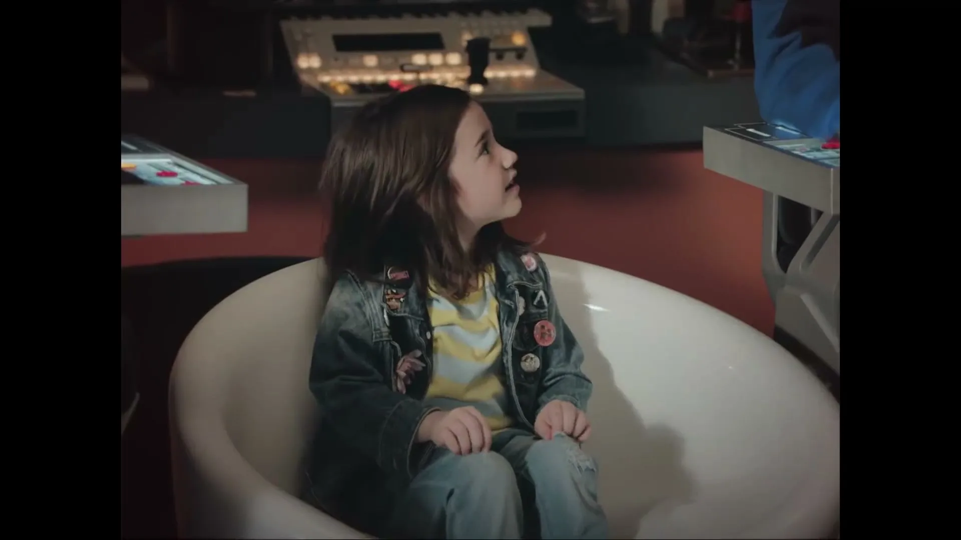 A young girl with long brown hair, wearing a denim jacket with pins, sits in a white chair in a dimly lit room with control panels, looking around curiously.