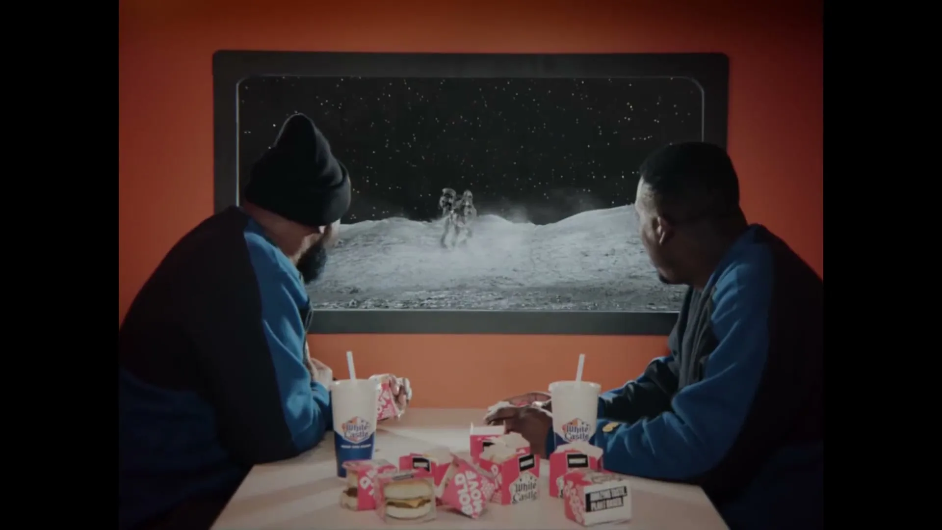Two men in blue and black tracksuits sit at a table, eating White Castle sliders and watching astronauts on the moon through a window.