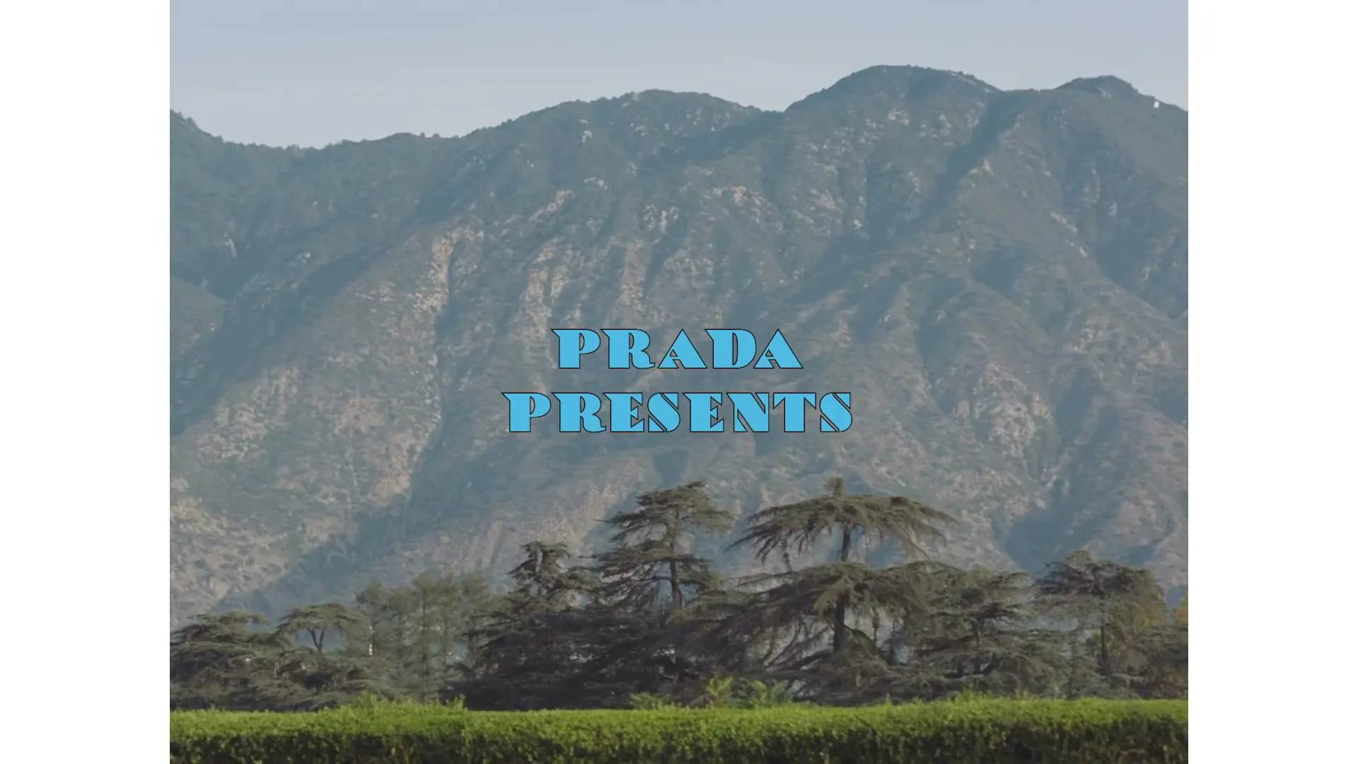 A wide shot of a mountain range with trees and a hedge in the foreground, with "PRADA PRESENTS" text overlaid.