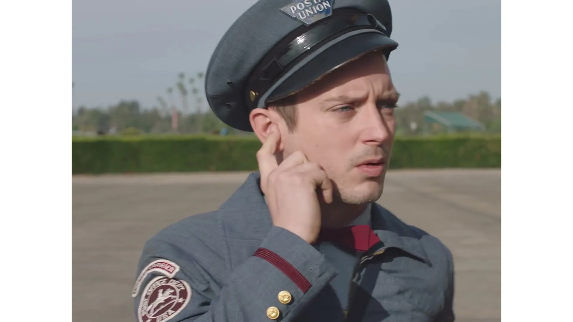 A male postman in a grey uniform and red bow tie looks around nervously in an empty outdoor parking lot during the day.