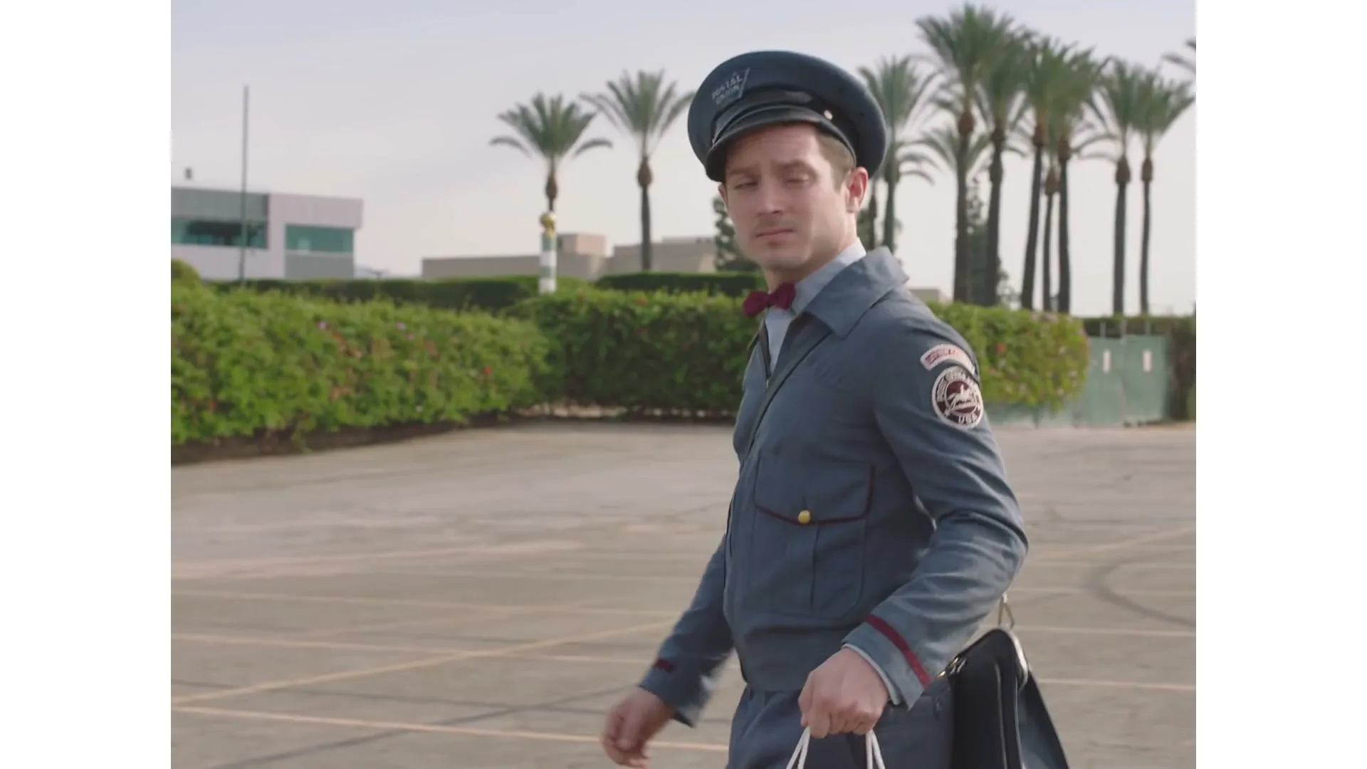 A man in a postman uniform walks across an empty parking lot, looking around suspiciously, with palm trees in the background.
