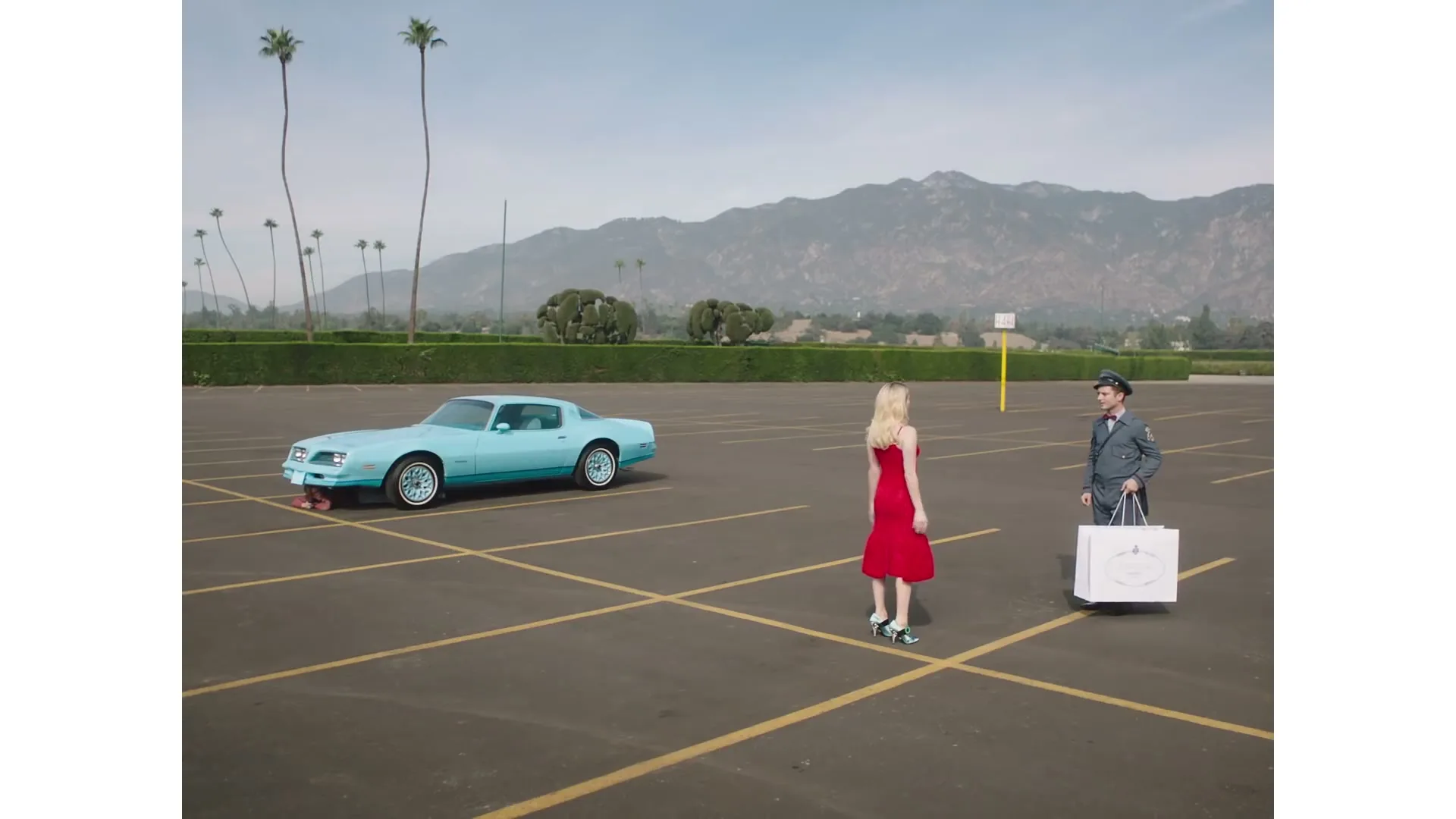 A woman in a red dress and a man in a suit stand in a vast, empty parking lot with a blue car, palm trees, and mountains in the background.