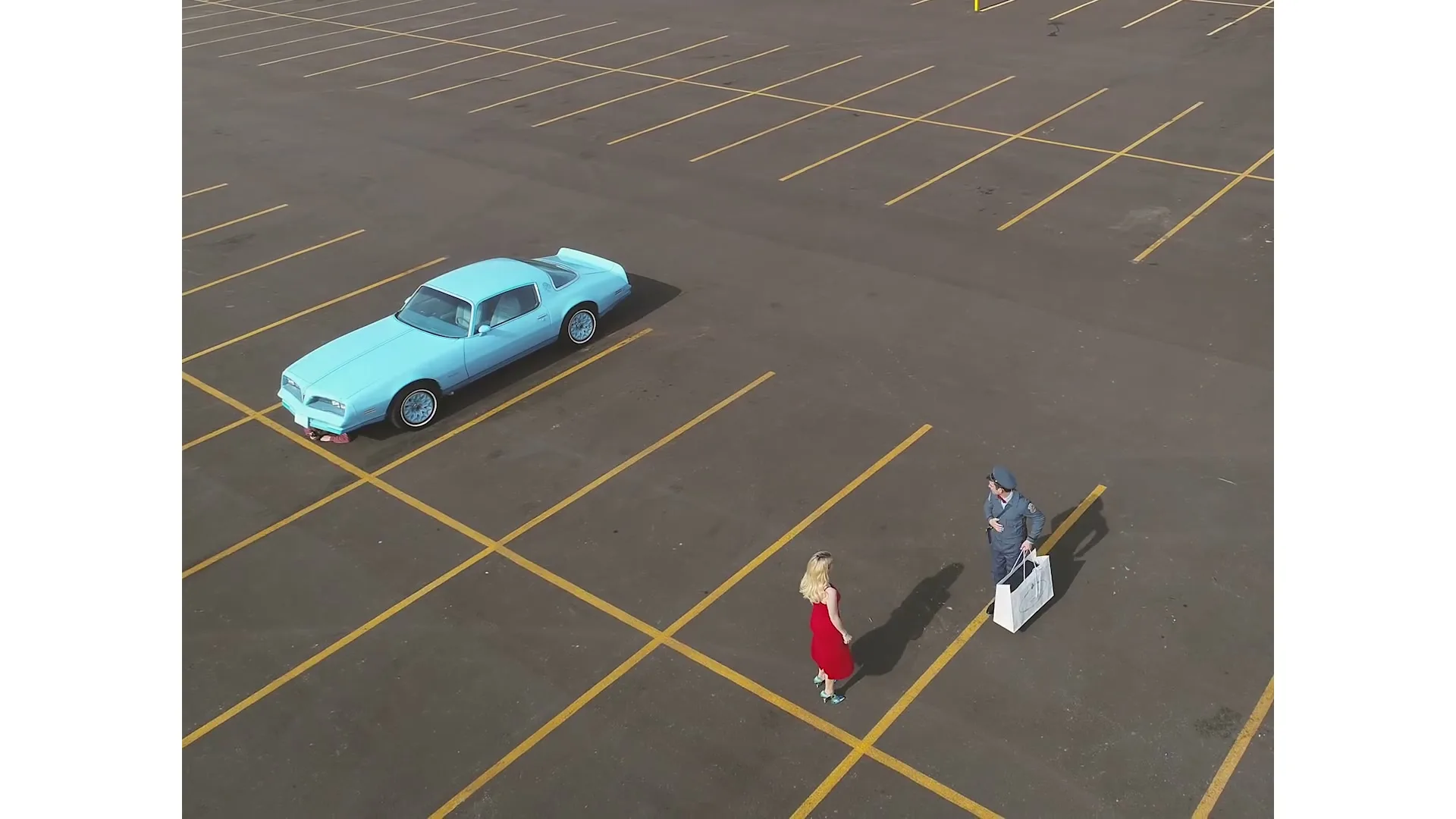A postman in uniform holds a white shopping bag while talking to a woman in a red dress in an empty parking lot with a blue car.