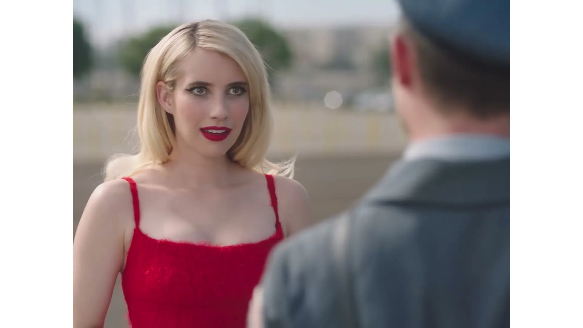 A blonde woman in a red dress smiles at a man in a uniform, who is seen from behind, outdoors on a sunny day.
