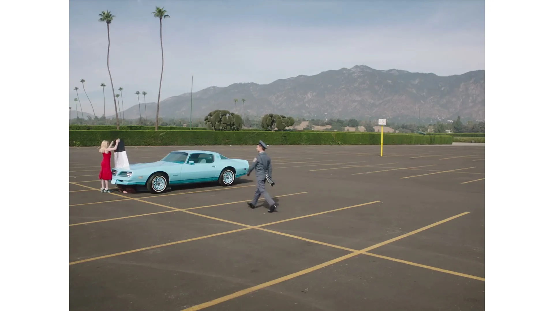 A woman in a red dress and a man in a uniform are standing next to a light blue car in a vast, empty parking lot with mountains in the background.