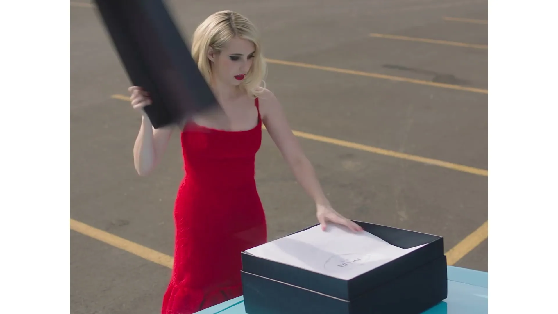 A blonde woman in a red dress opens a black box with a white tissue paper inside, revealing a Prada logo, in an outdoor parking lot.