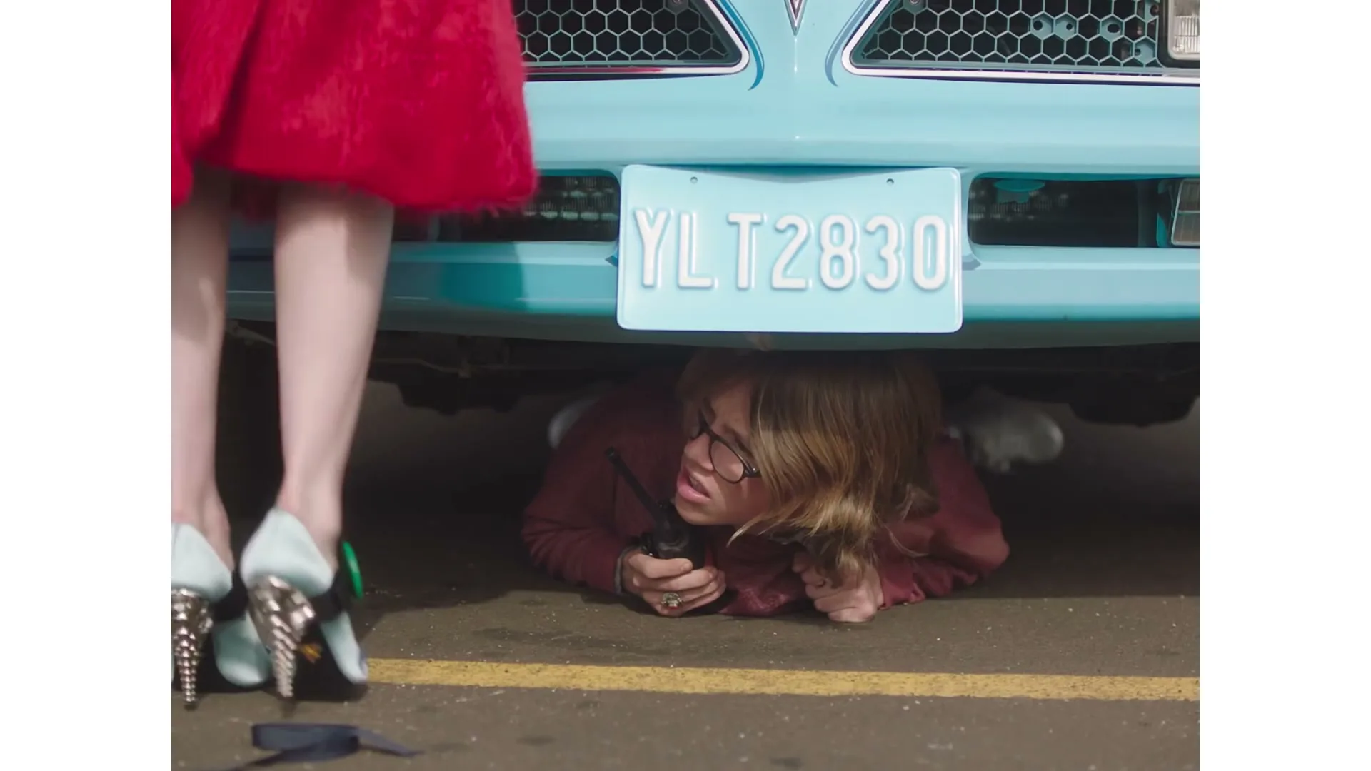 A woman in a red dress and high heels stands over another woman who is lying on the ground under a light blue car, holding a walkie-talkie.