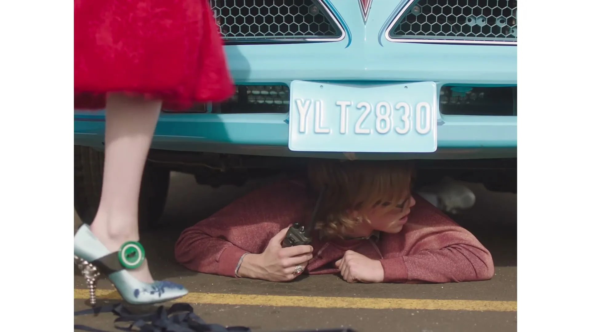A woman in a red dress and blue heels stands over another woman lying under a light blue car, who is holding a walkie-talkie.