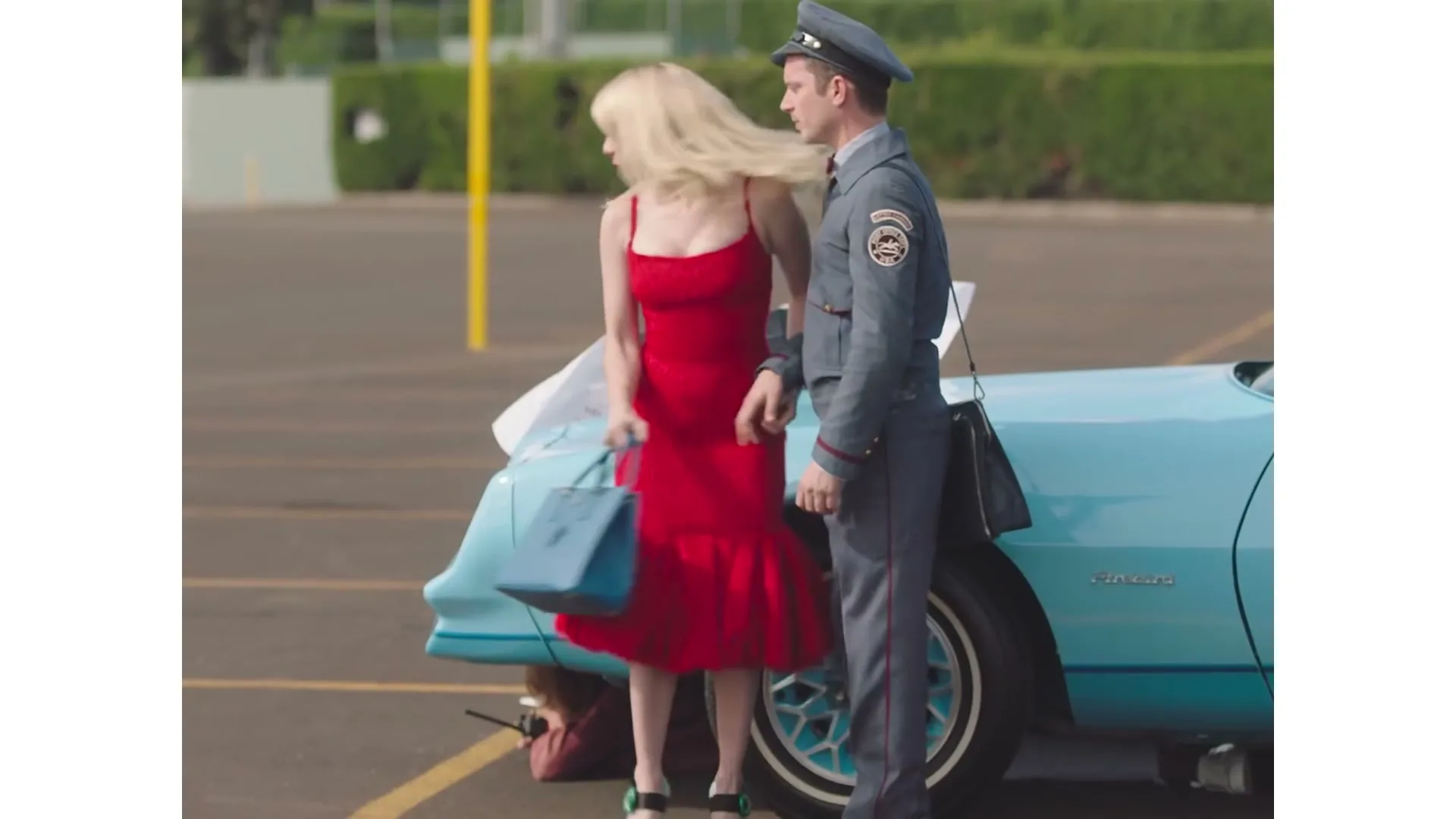 A blonde woman in a red dress and a man in a postman uniform stand next to a light blue car in a parking lot.