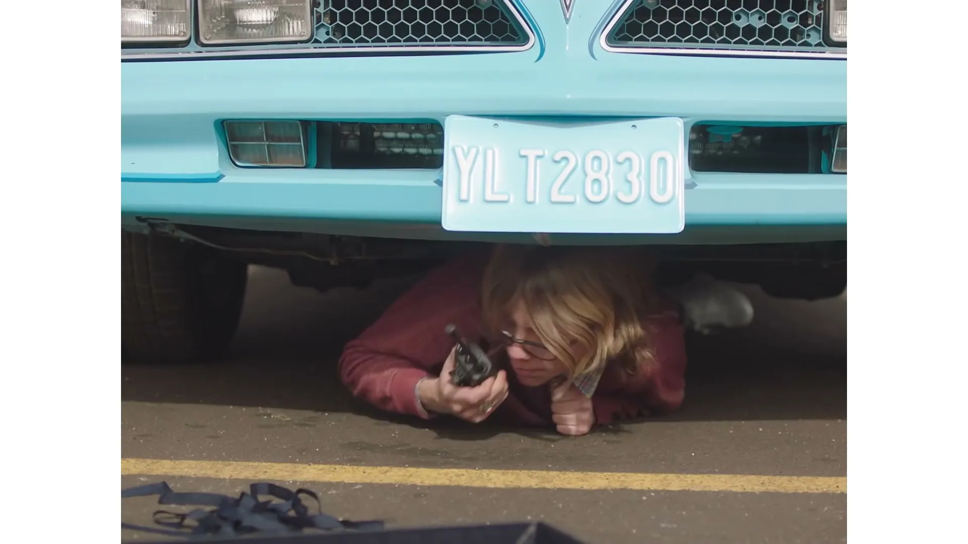 A person with blonde hair and glasses lies on the ground under a light blue car, holding a walkie-talkie and looking up.
