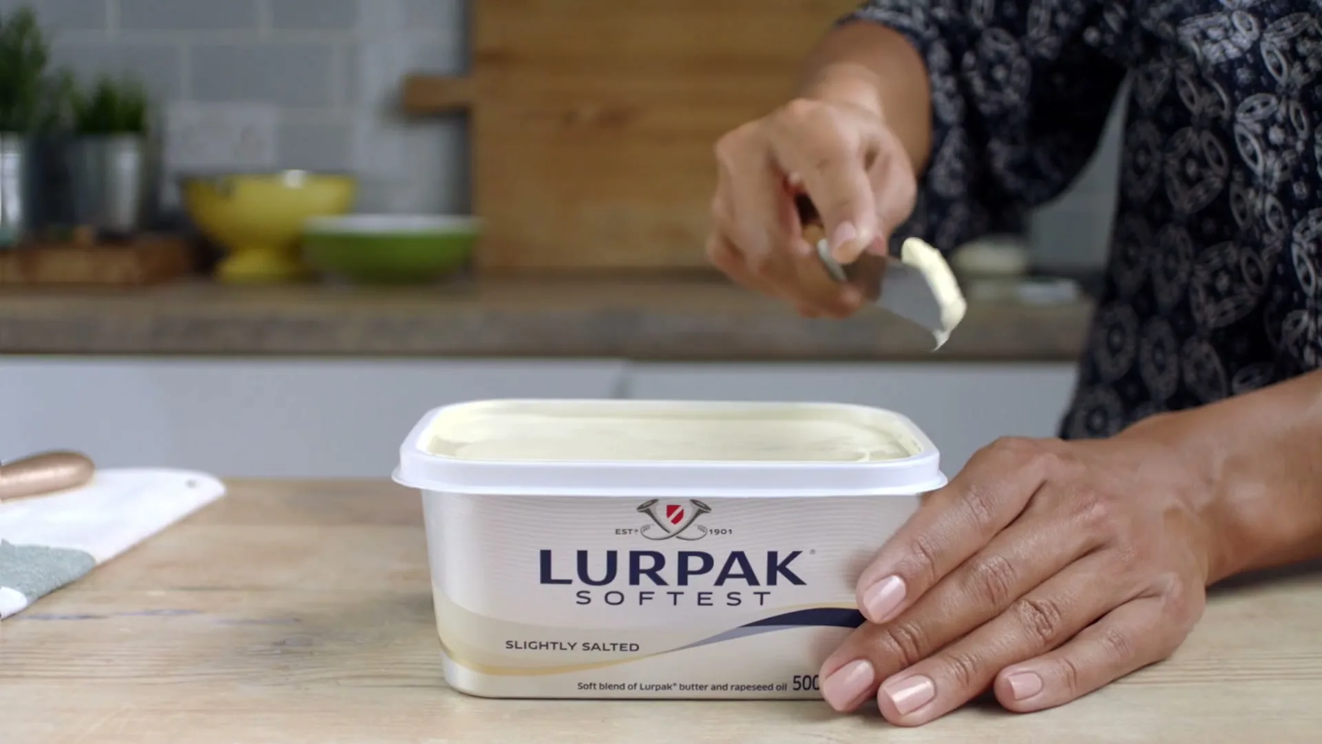 A woman's hands are seen scooping soft butter from a Lurpak tub with a knife on a wooden kitchen counter.
