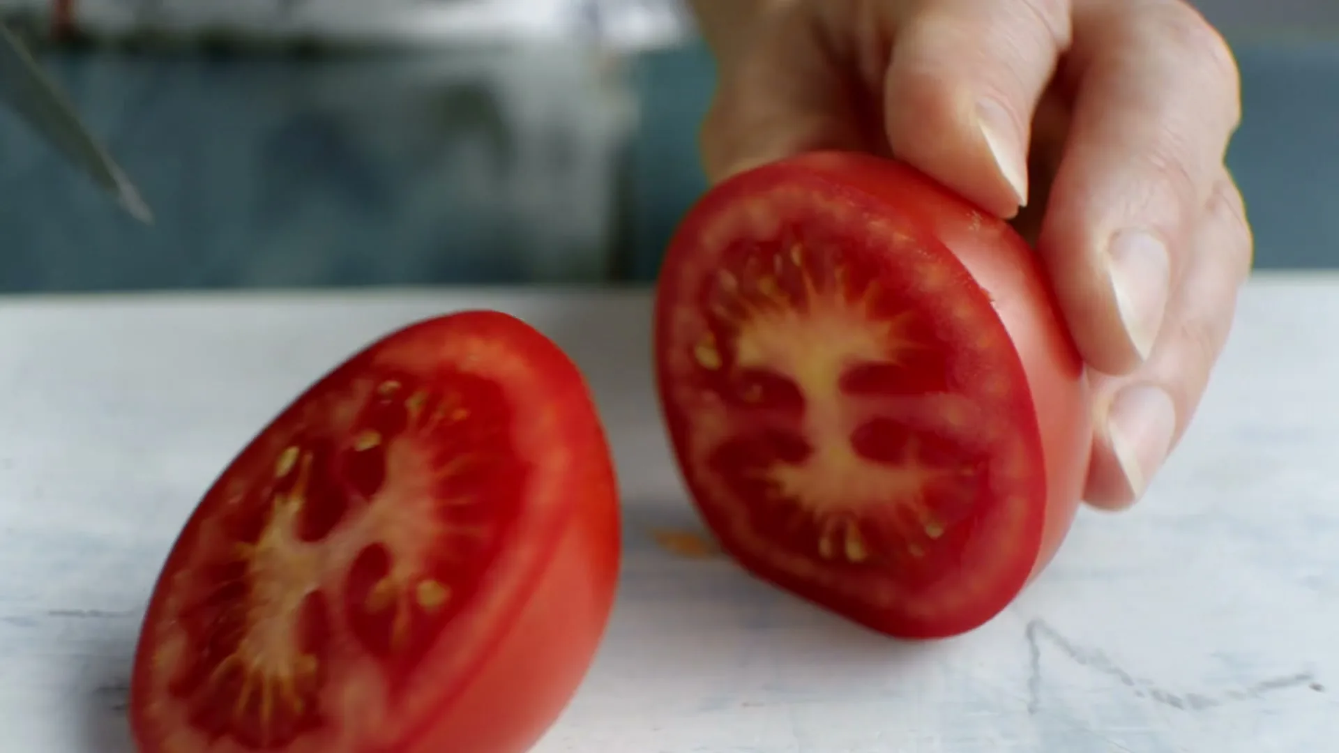 A close-up shot shows a person's hand holding a red tomato on a white cutting board, as a knife slices through it, revealing the juicy interior.