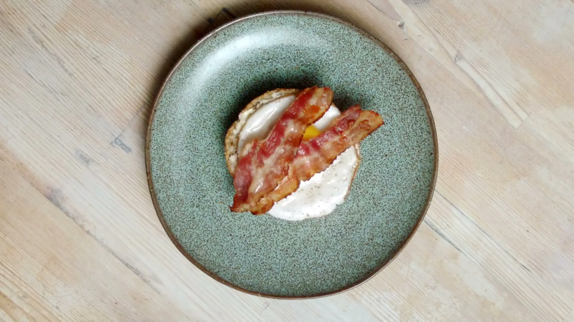 A top-down shot shows a bread roll being assembled on a green plate, with bacon and a fried egg added, then topped with the other half of the roll.