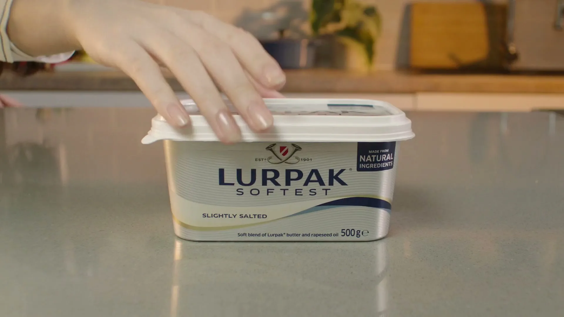 A woman's hand reaches down to open a tub of Lurpak Softest butter on a kitchen counter.