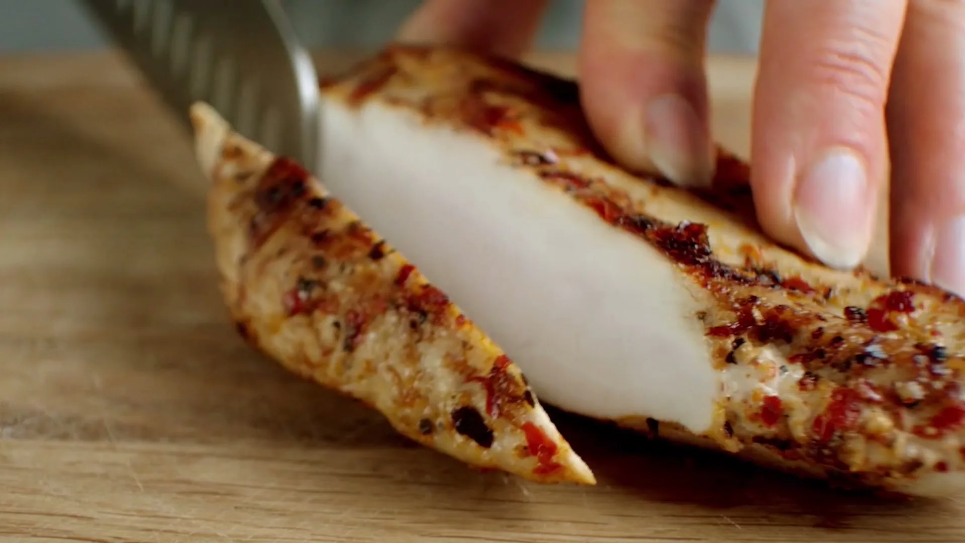 A close-up shot shows a person's hand holding a knife, slicing a piece of cooked, seasoned chicken on a wooden cutting board.