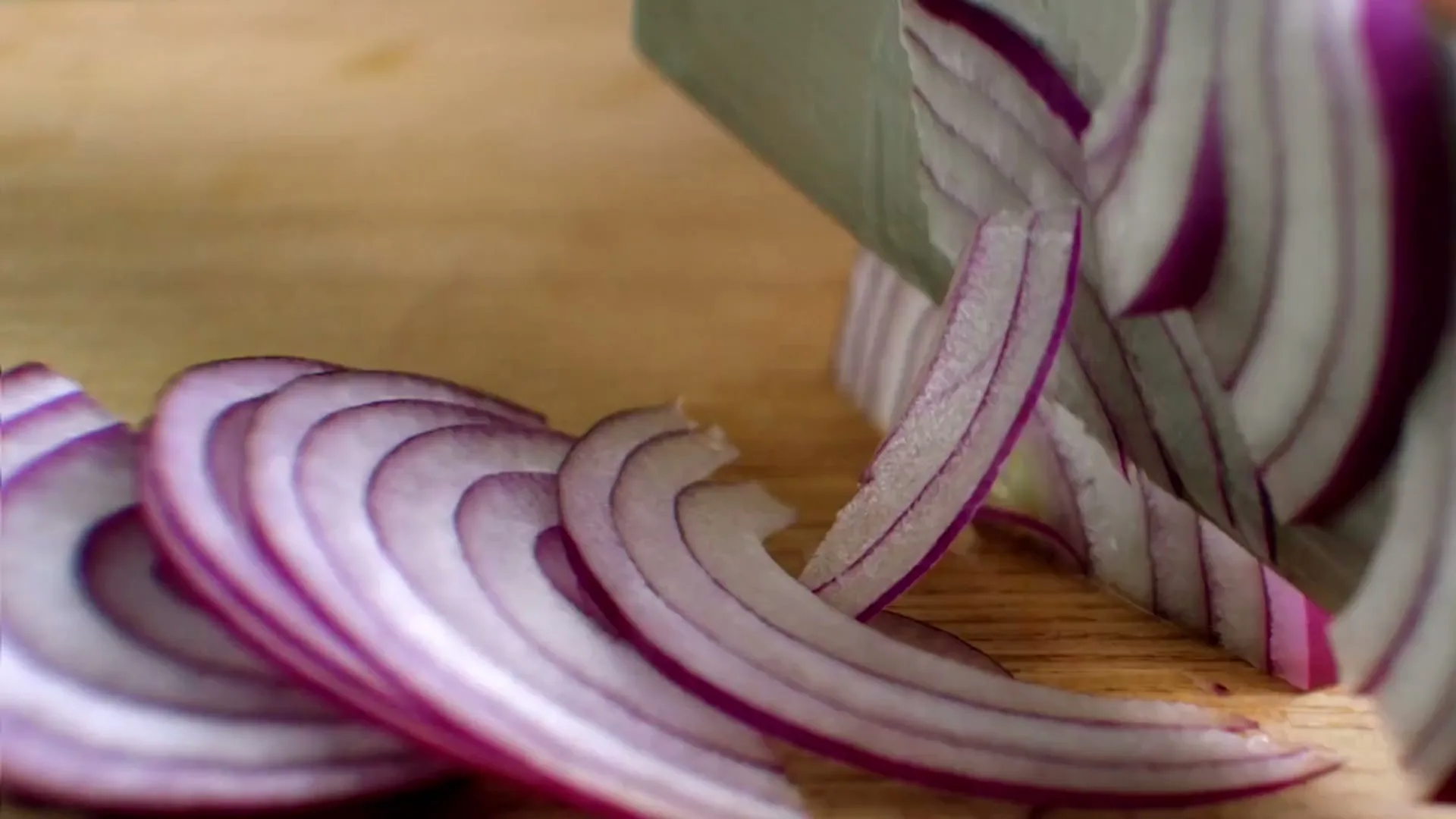 A close-up shot shows a knife slicing thin, purple rings from a red onion on a light wooden cutting board.