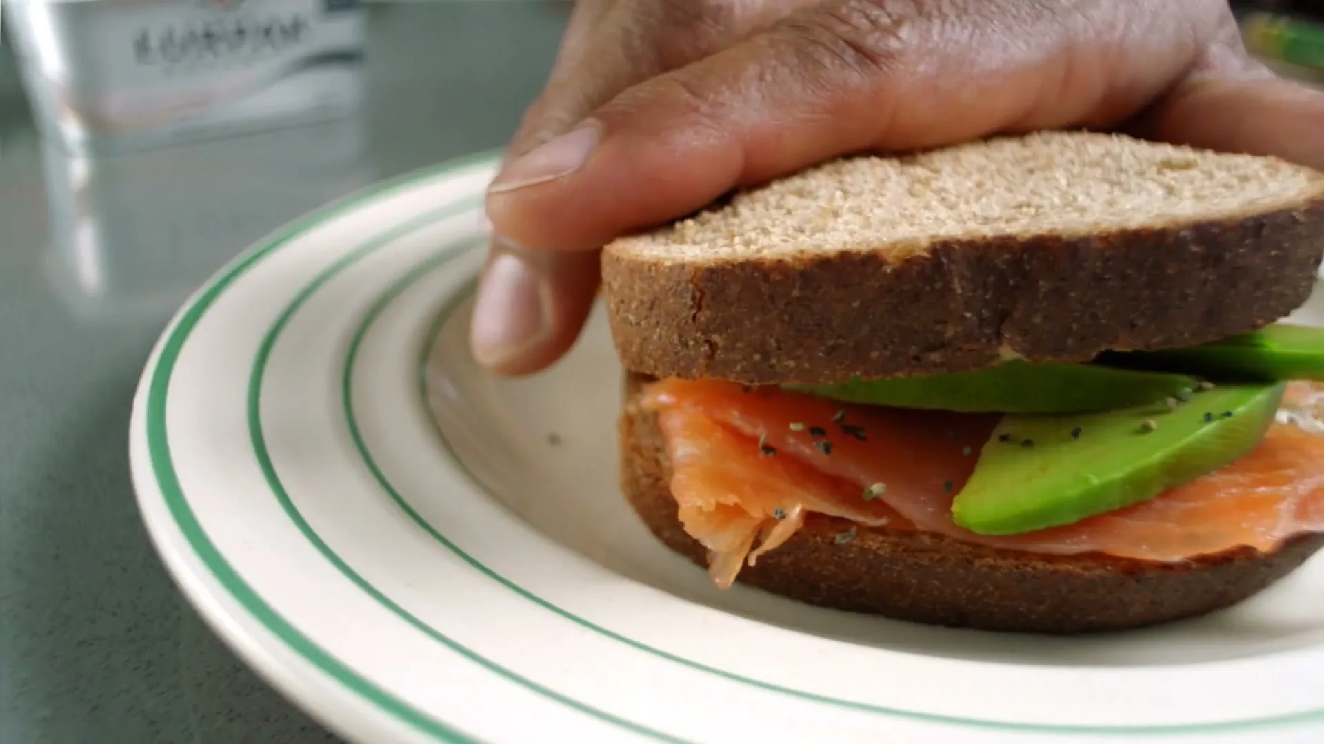 A person's hand holds the top slice of a salmon and avocado sandwich on a white plate with green stripes, in a kitchen.