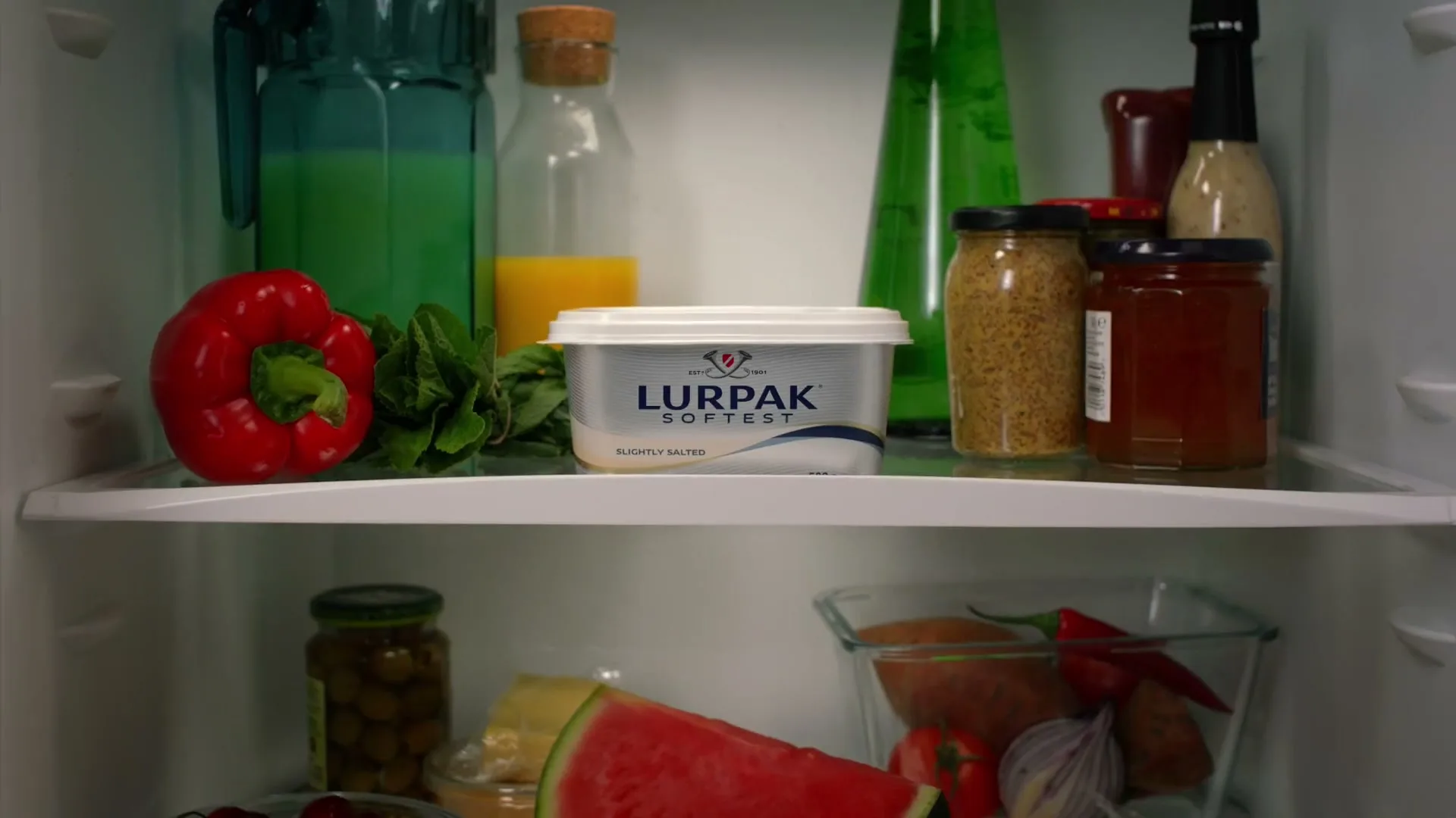 A tub of Lurpak Softest butter sits on a shelf in a refrigerator, surrounded by various fresh foods and condiments.