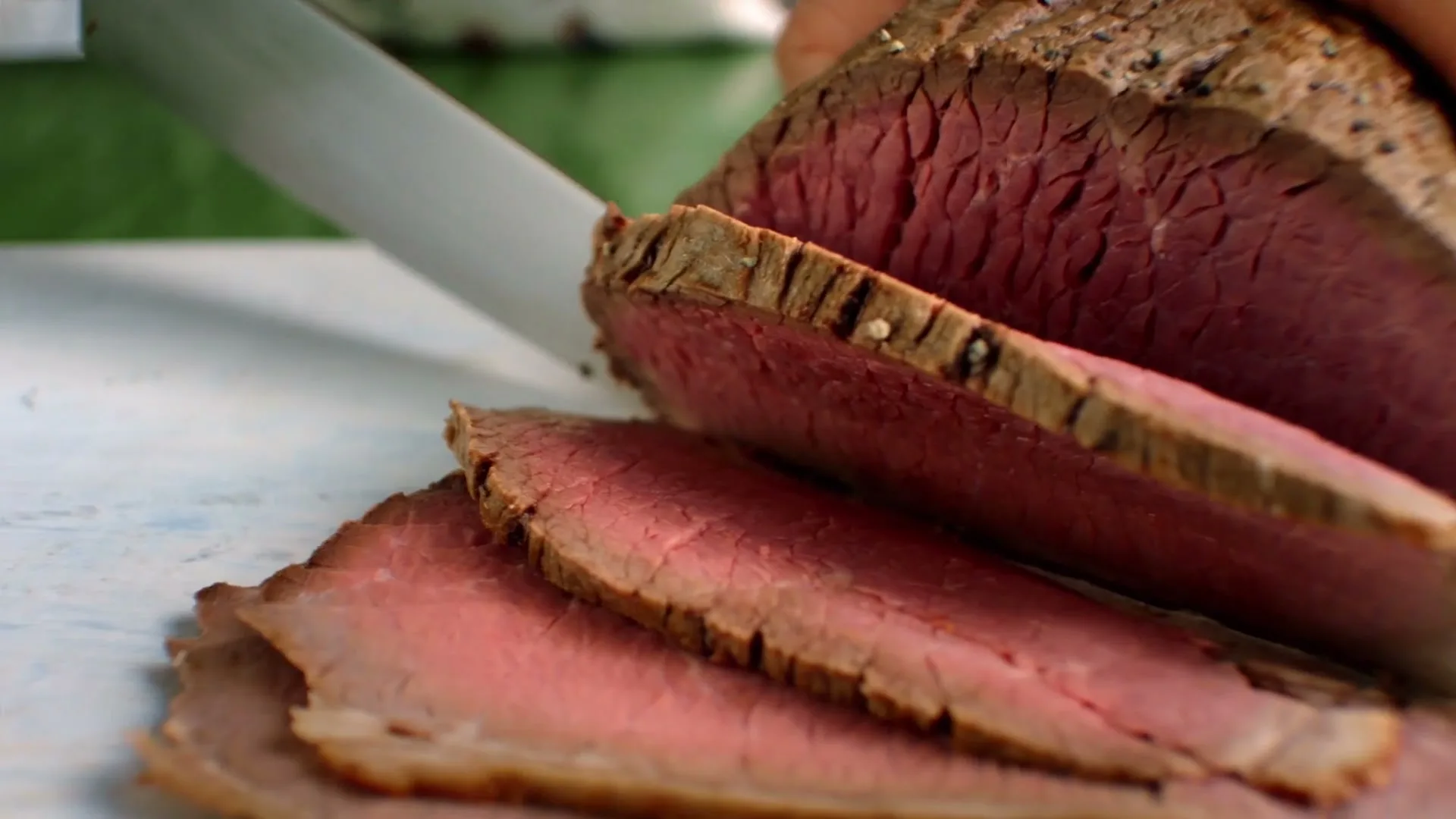 A chef's hands slice a perfectly cooked, medium-rare roast beef on a white cutting board in a kitchen.