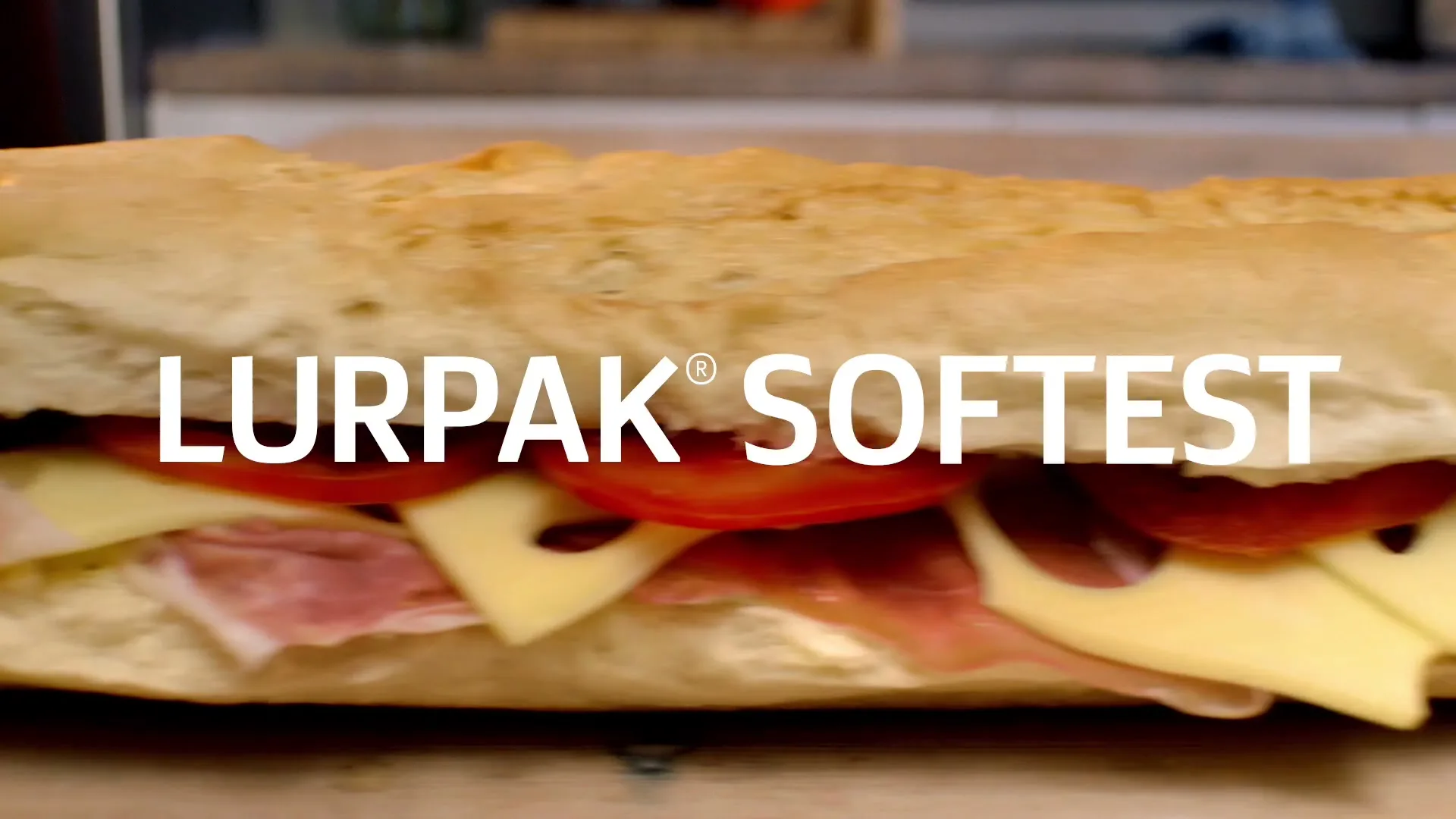 A close-up shot of a delicious-looking sandwich with ham, cheese, and tomatoes, with the text "LURPAK
SOFTES" overlaid on top.