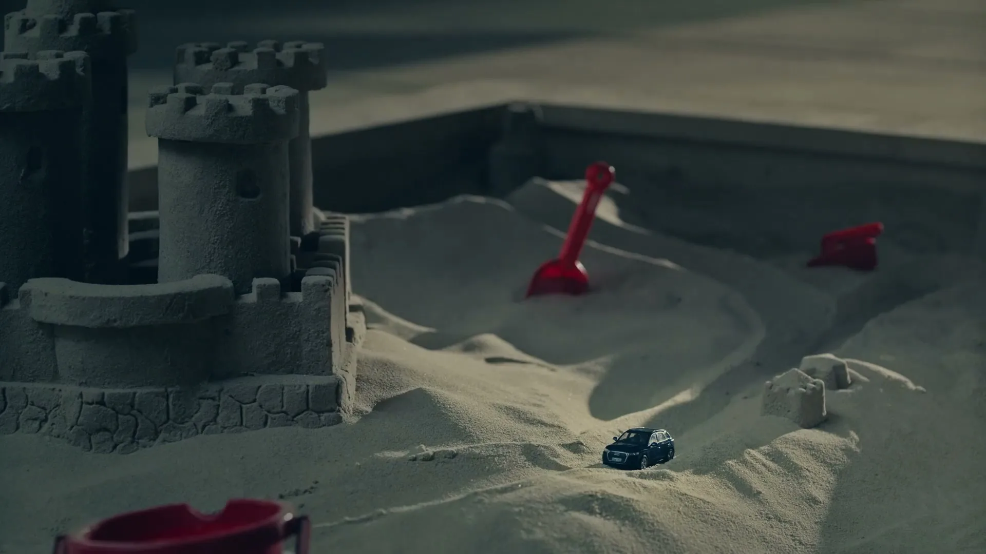 A toy car sits in a sandbox next to a sandcastle and red plastic toys, under dim lighting.