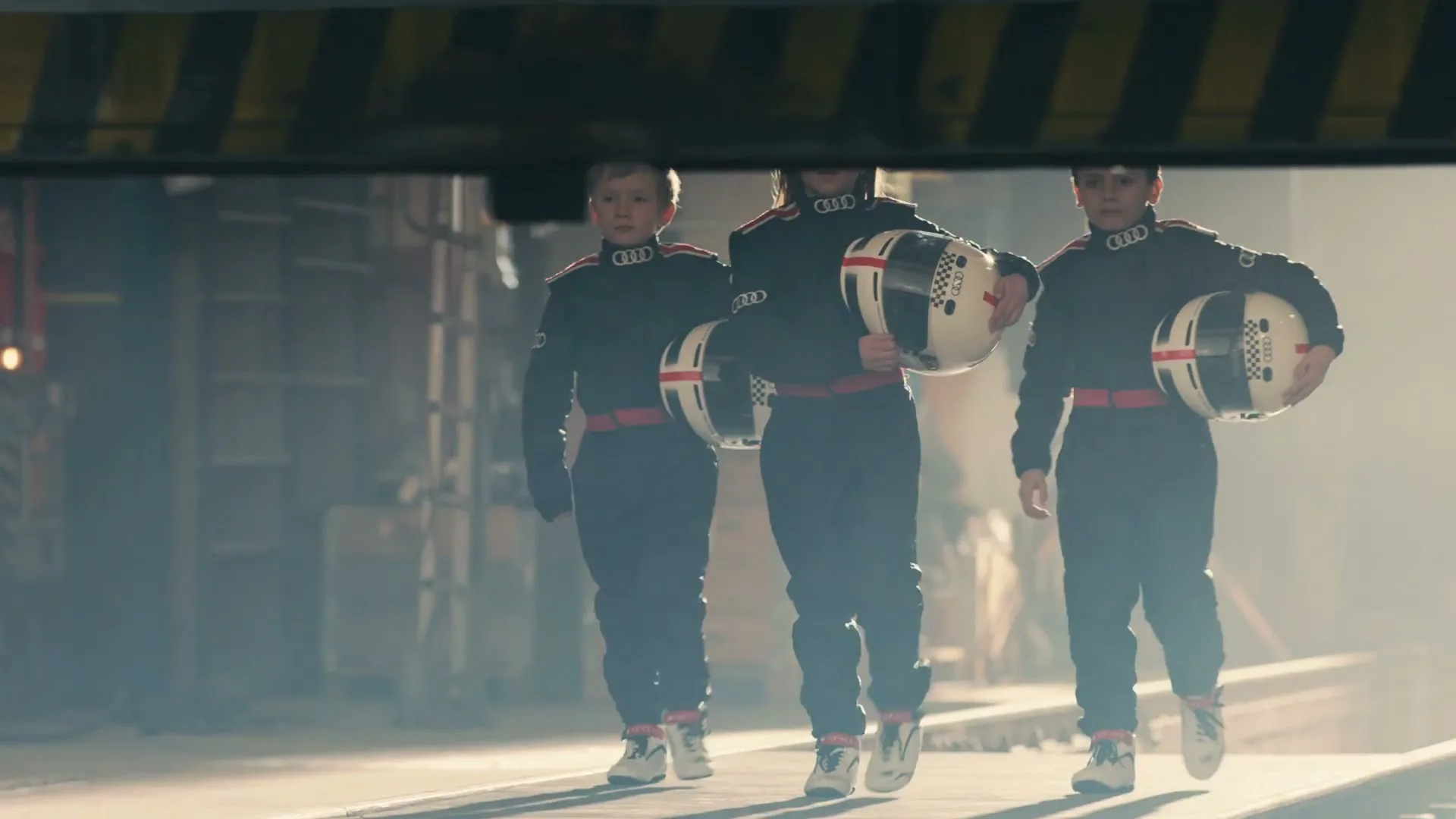 Three children in racing suits and helmets walk towards the camera in a dusty, industrial setting, with an overhead beam in the foreground.