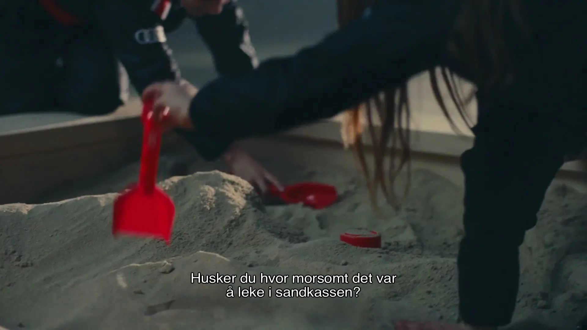 Two children in dark clothing play in a sandbox with red plastic toys, digging in the sand.