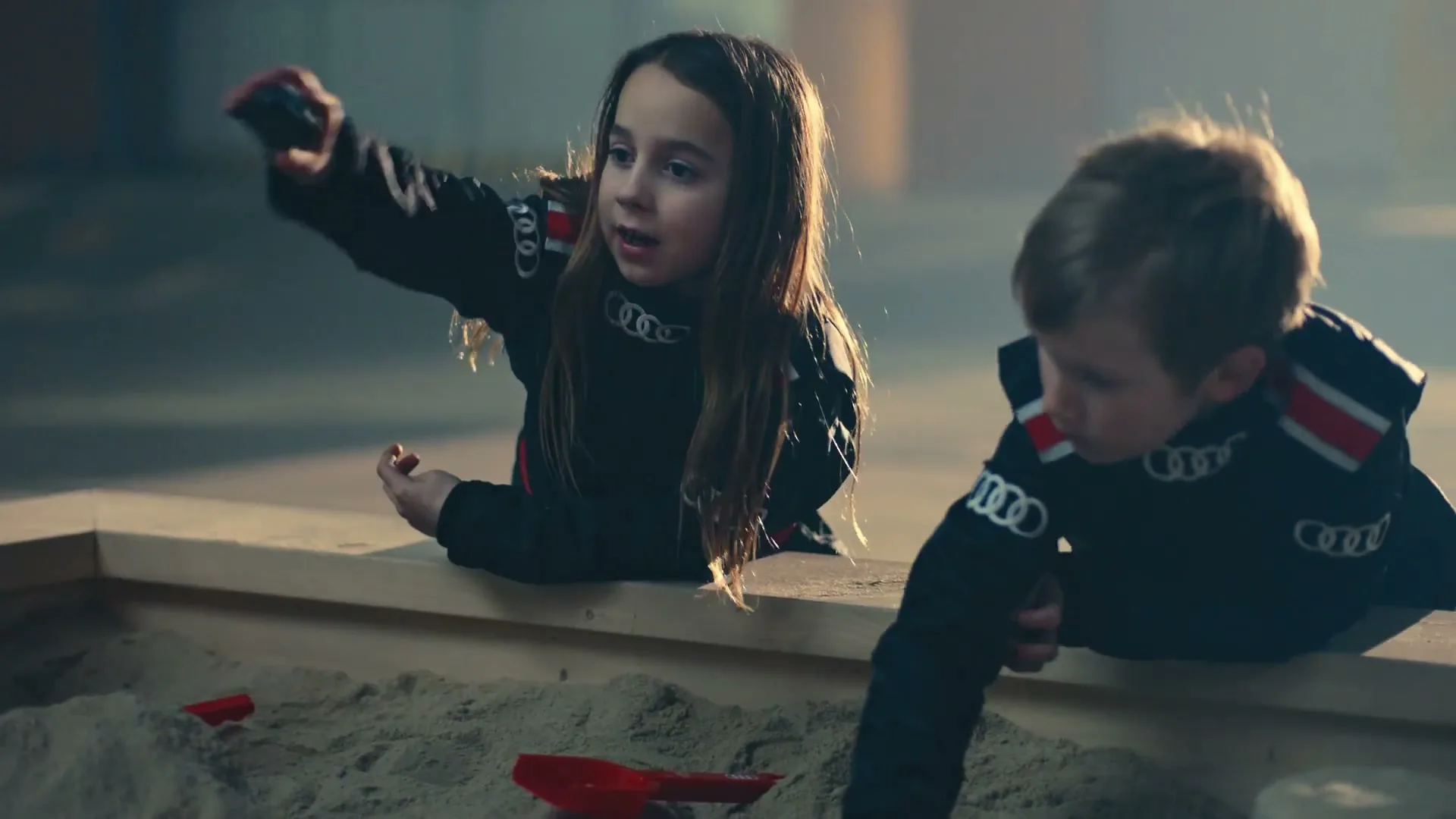 Two children, a girl and a boy, wearing black racing suits with Audi logos, play in a sandbox in a dimly lit, industrial-looking interior.