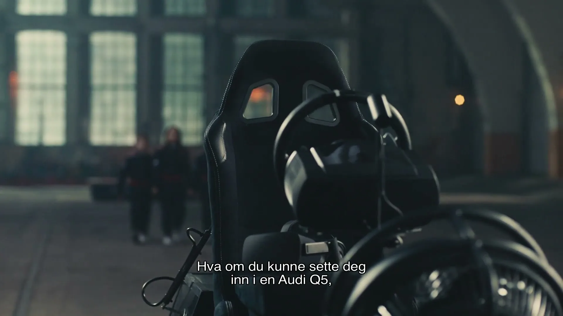 Three children in racing suits walk towards a racing simulator in a large, industrial warehouse.