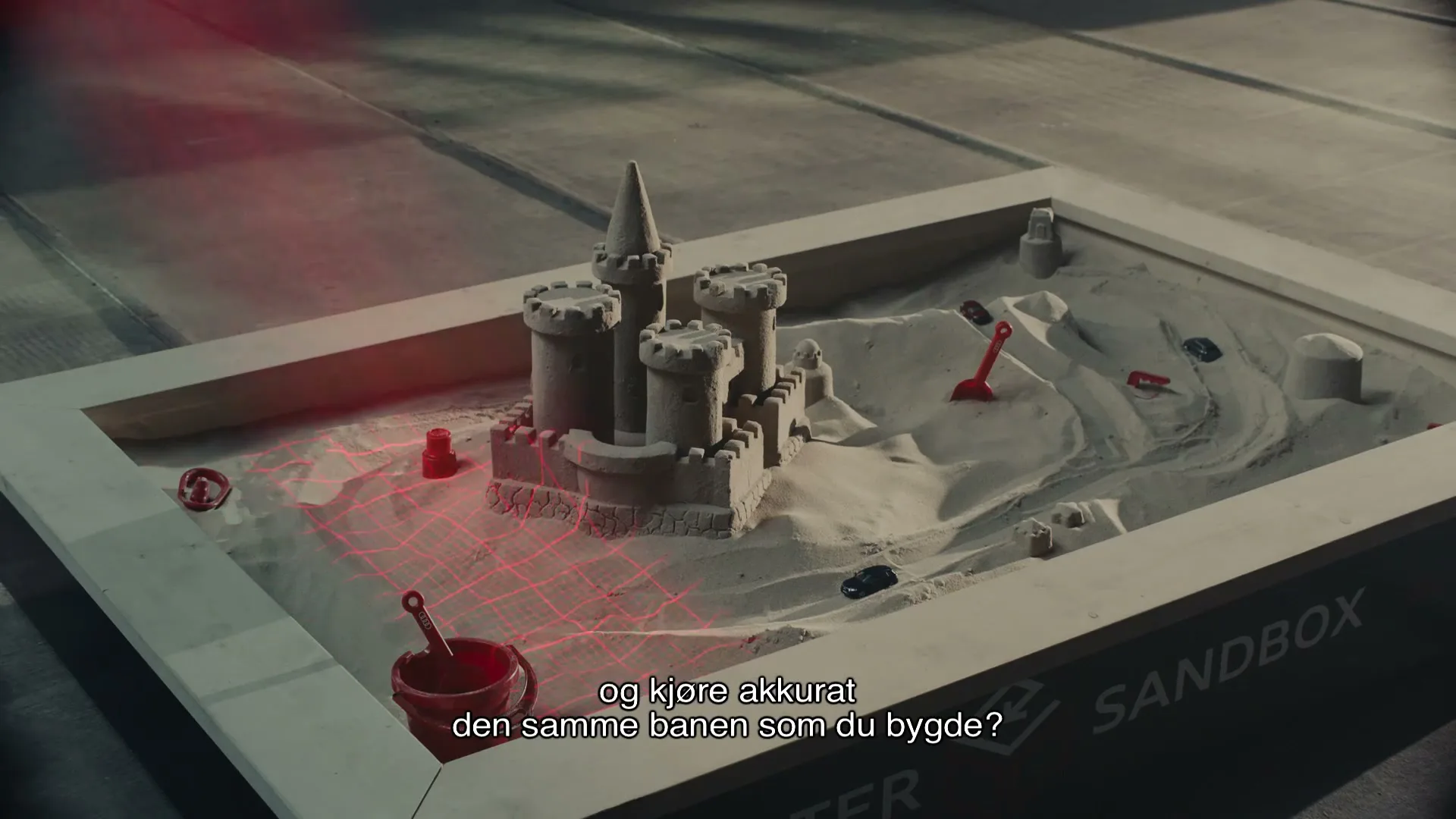 A sandcastle in a sandbox with red toy shovels and buckets, as red laser lines trace paths around it, in an interior setting.