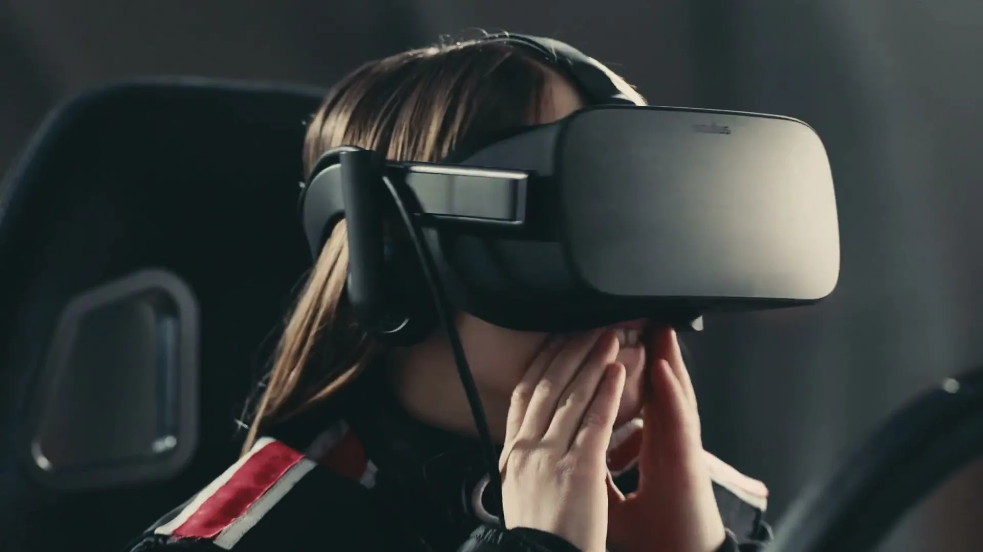 A young woman with long brown hair wears a VR headset and a racing suit, covering her mouth with her hands in a dark interior.
