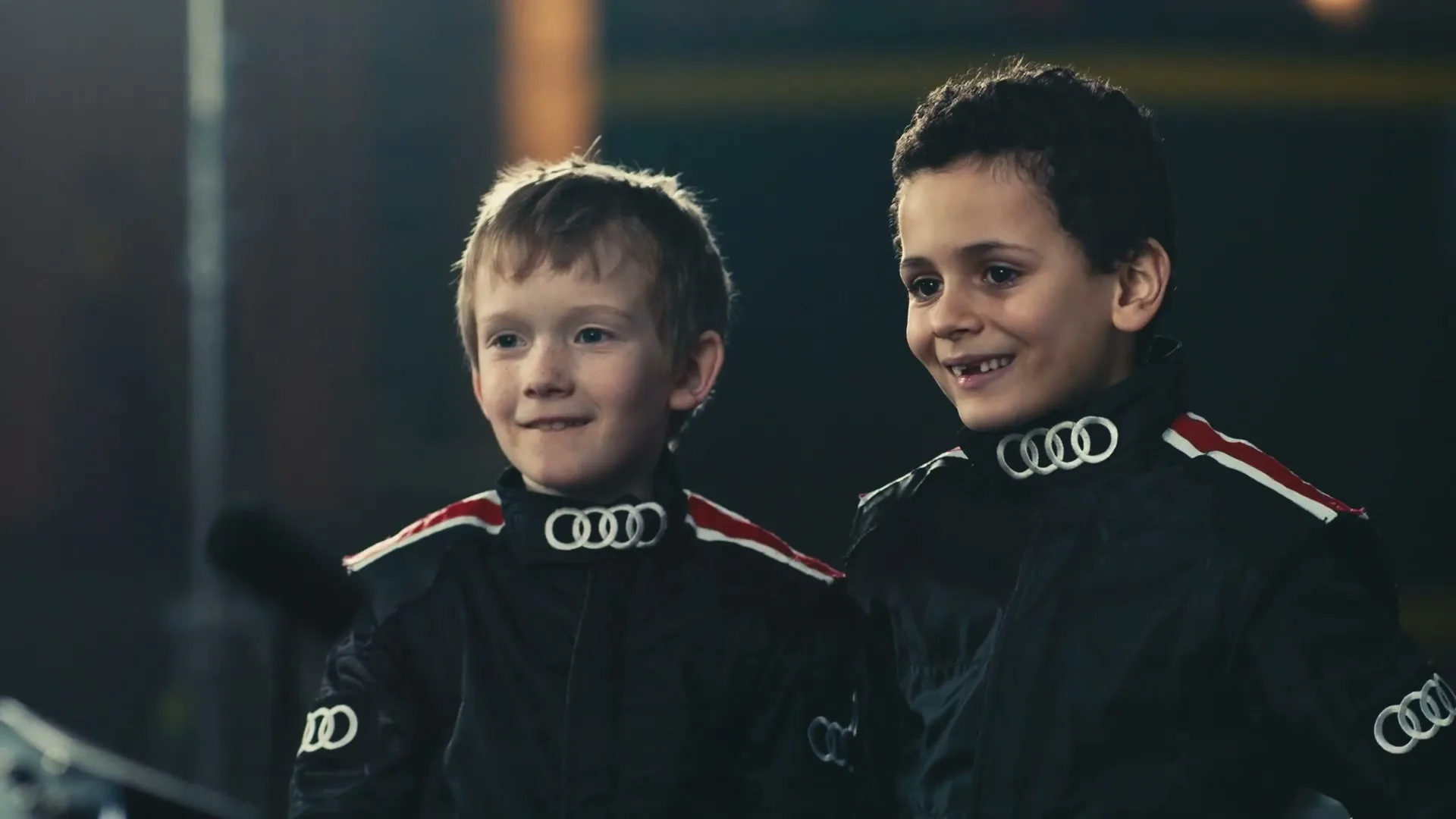 Two young boys in black racing suits with Audi logos smile and look forward in an indoor setting.