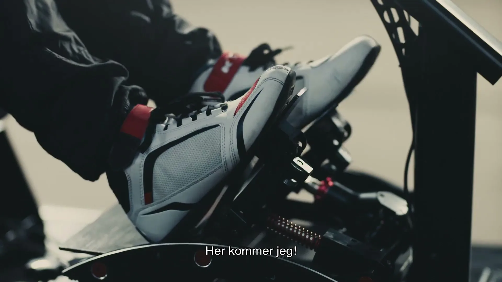 A person's feet in white racing shoes are shown pressing pedals in a vehicle, with the text "Her kommer jeg!" on screen.