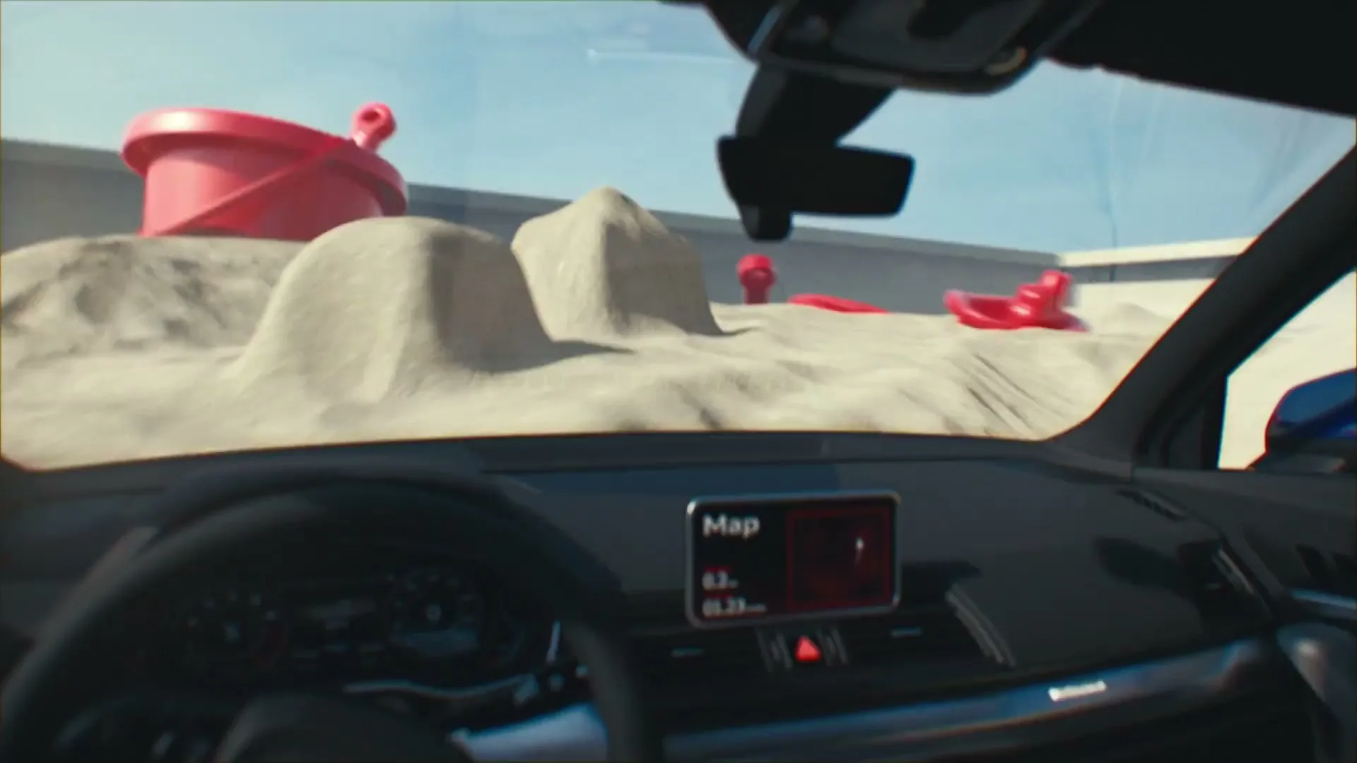 The interior of an Audi car is shown, looking out the windshield at a sandy landscape with red toy shovels and buckets.