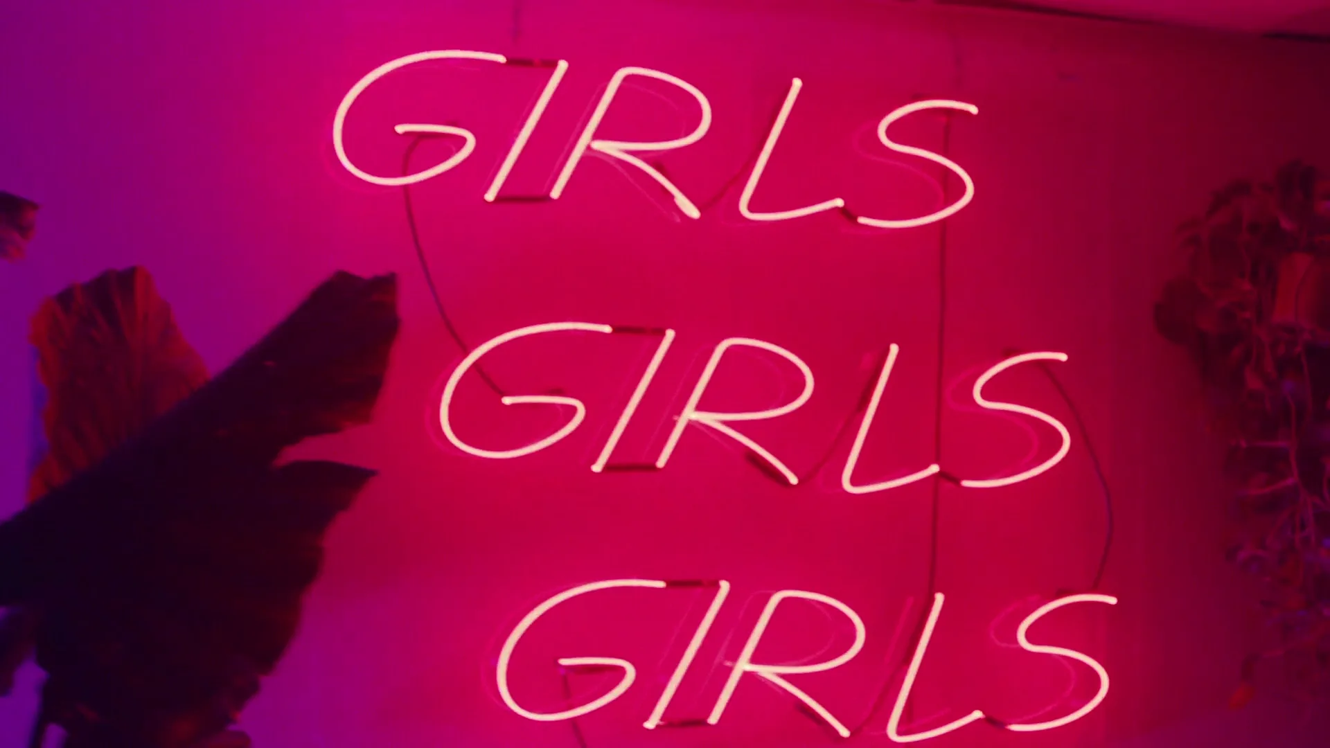 A neon sign with the word "GIRLS" repeated three times glows brightly on a pink wall in an interior setting.