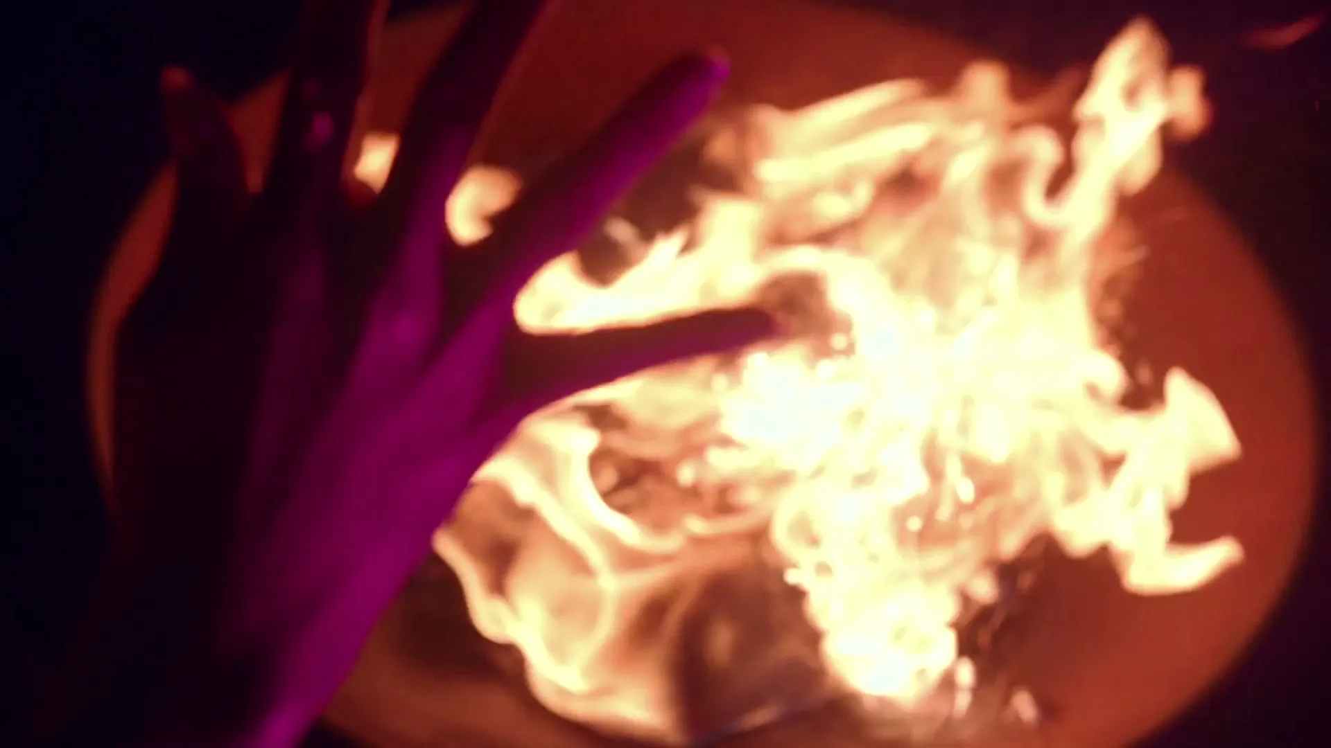 A woman's hand with purple light on it is shown above a fire pit with bright orange flames and sparks flying upwards in a dark setting.