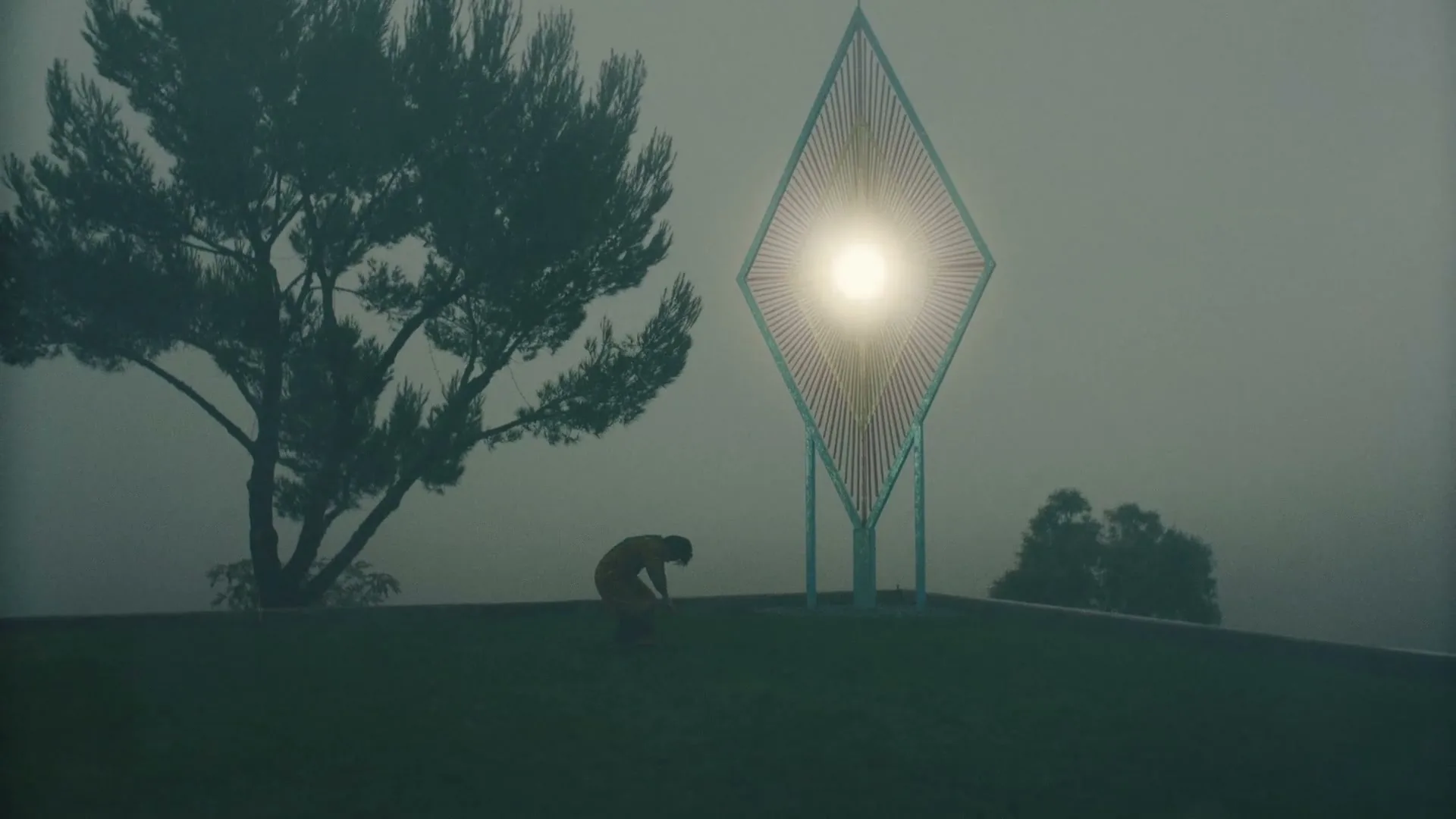 A person in a yellow jumpsuit is seen from a distance, bending over on a grassy hill next to a large, glowing diamond-shaped sculpture on a foggy day.