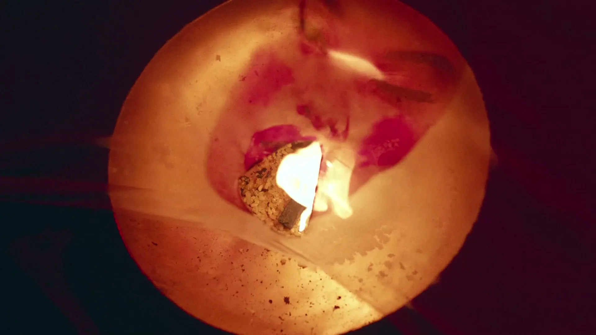 A woman's face is seen through a translucent, circular object with a small fire burning inside, creating a surreal and intense visual.