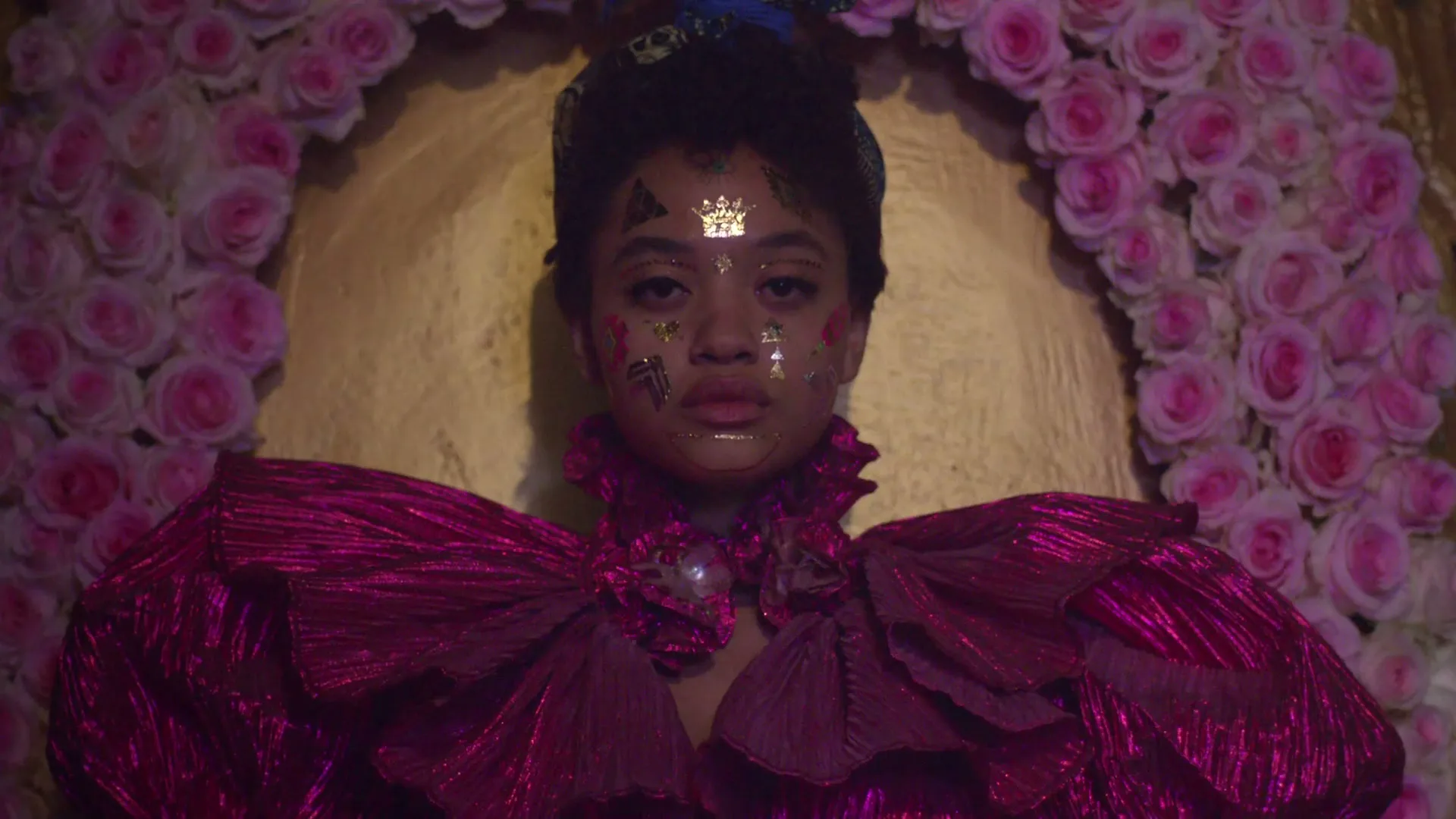 A young woman with gold face decals and a voluminous magenta dress stares directly at the camera, framed by a pink rose arch.