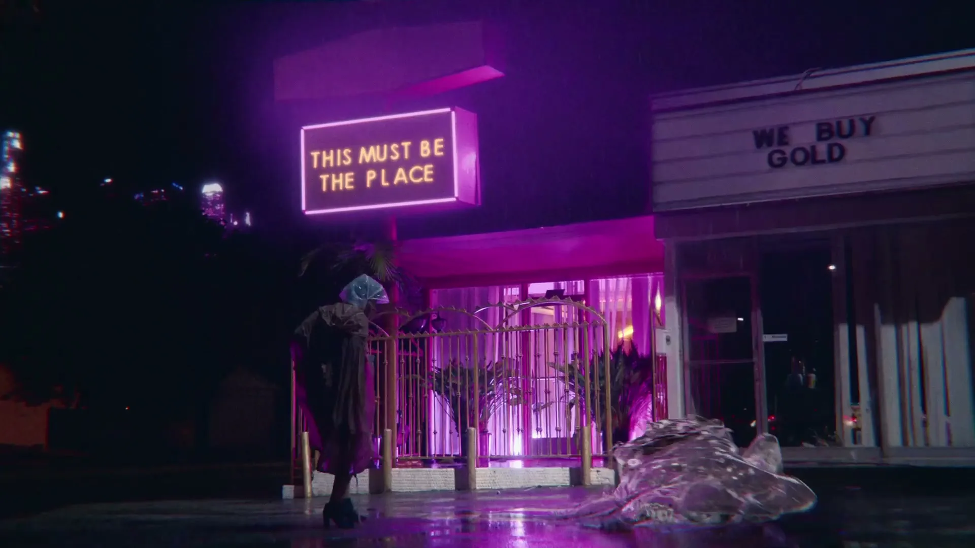 A person in a trench coat and blue head covering stands on a wet street at night, looking at a neon sign that reads "THIS MUST BE THE PLACE" next to a "WE BUY GOLD" store.
