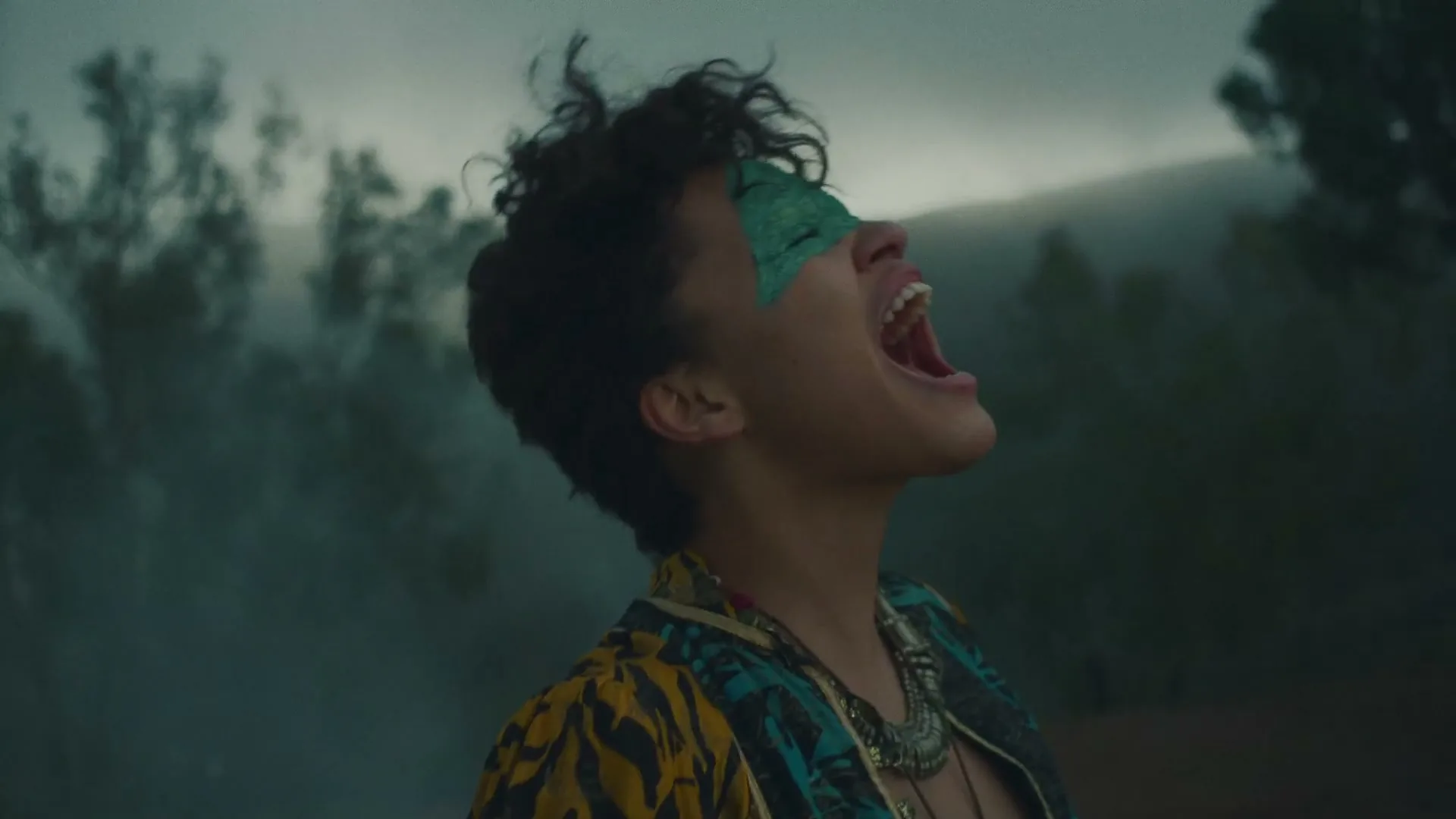 A woman with short curly hair and green face paint on her eyes screams with her head tilted back in a smoky, misty forest.