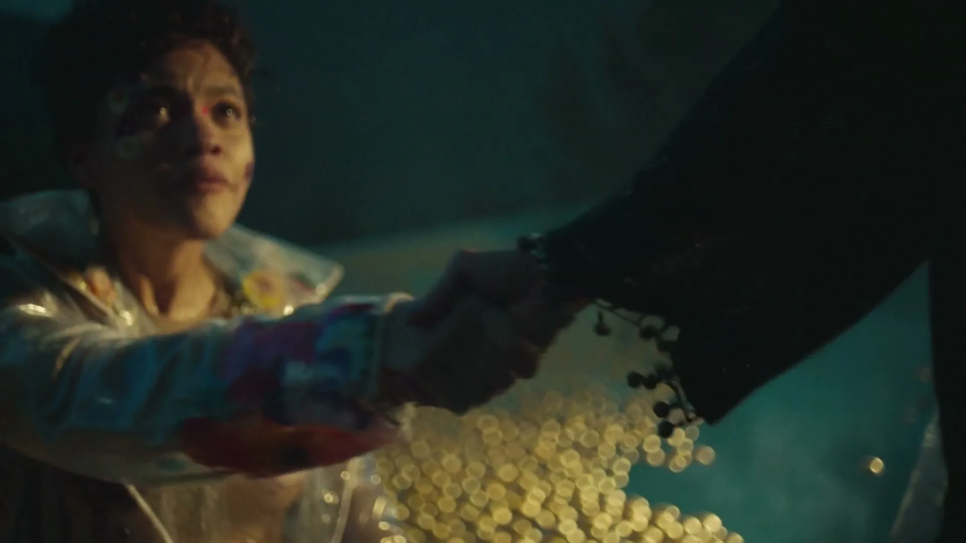 A young woman with painted face and clear raincoat reaches out to hold hands with a person in a dark, bell-sleeved top, as golden beads fall between them.
