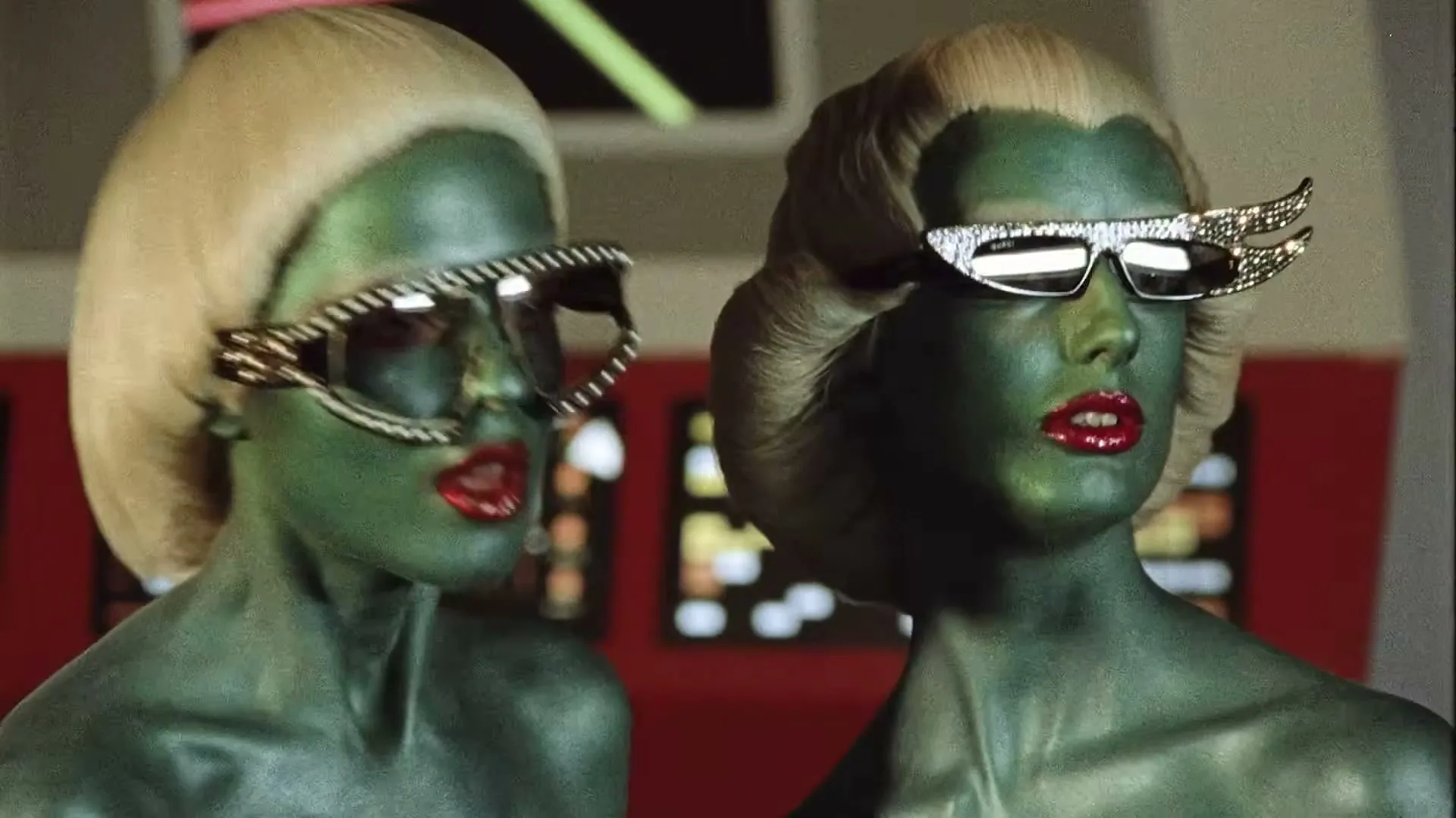 Two green-skinned alien women with blonde wigs and elaborate sunglasses look around a spaceship bridge.