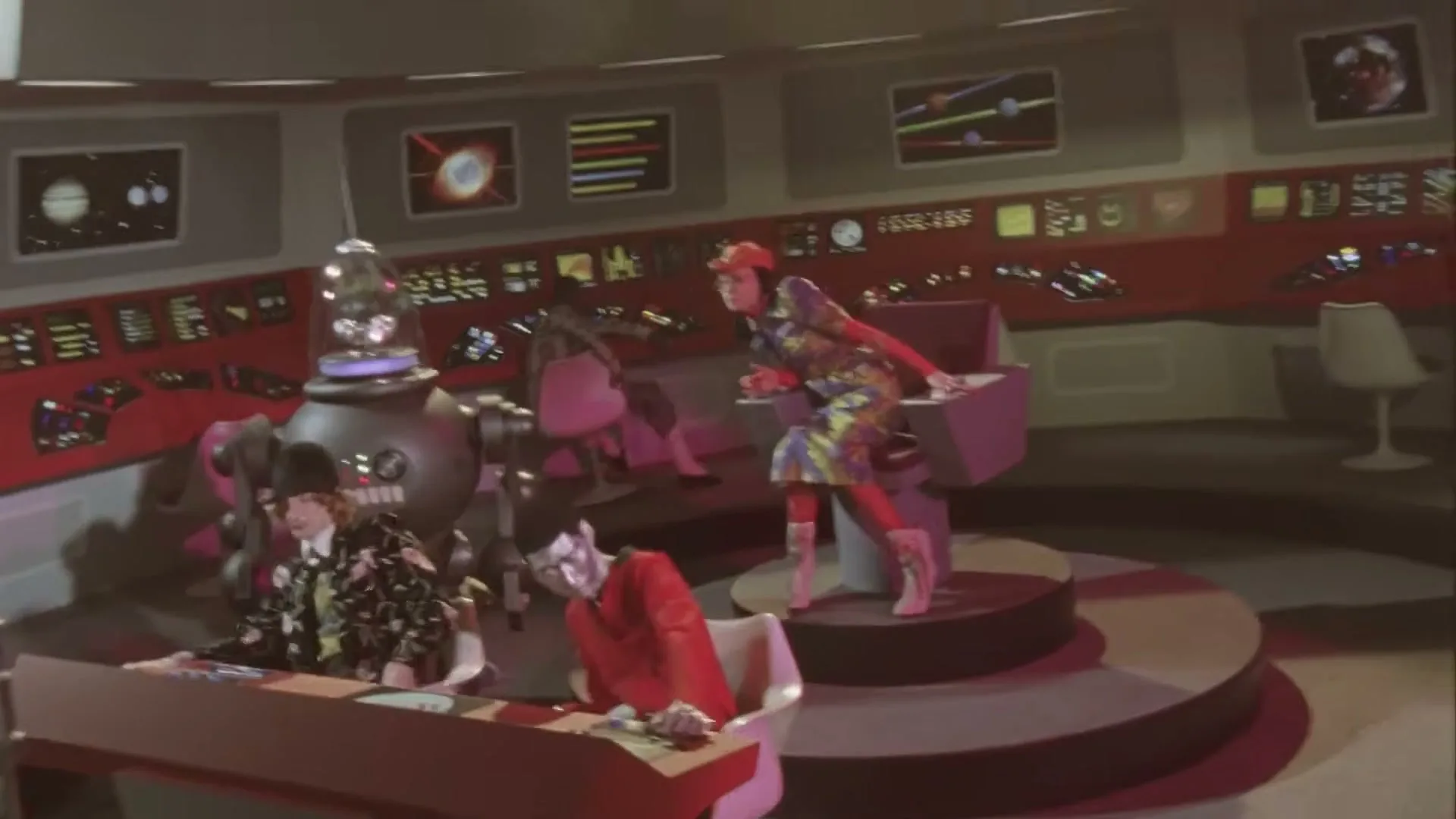 Three people in colorful, retro-futuristic outfits are in a spaceship control room, with one person in red falling out of their chair.