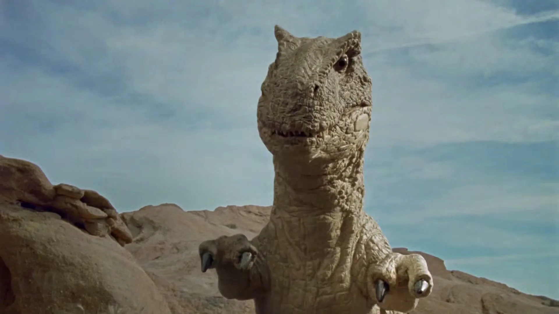 A dinosaur, resembling a Tyrannosaurus Rex, stands in a desert landscape, roaring with its mouth wide open under a cloudy sky.